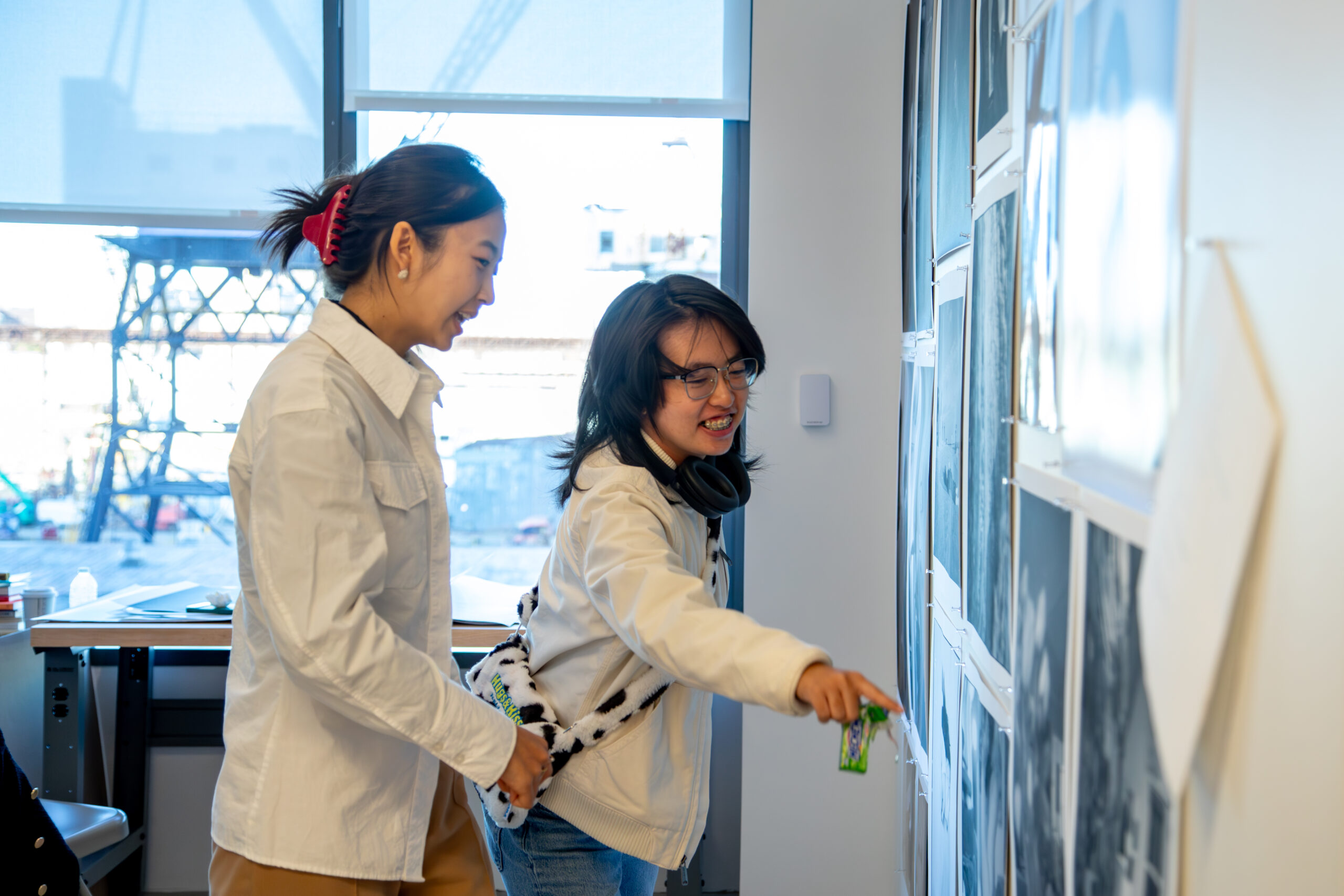 Two people smiling and engaging with a wall display of black-and-white photographs in an art studio. One person is pointing at a photo while holding a small green object, and the other stands beside them, watching with interest. Both are dressed casually in light-colored jackets, and one has headphones around their neck. A large window in the background reveals an industrial outdoor scene with cranes, adding natural light to the space.