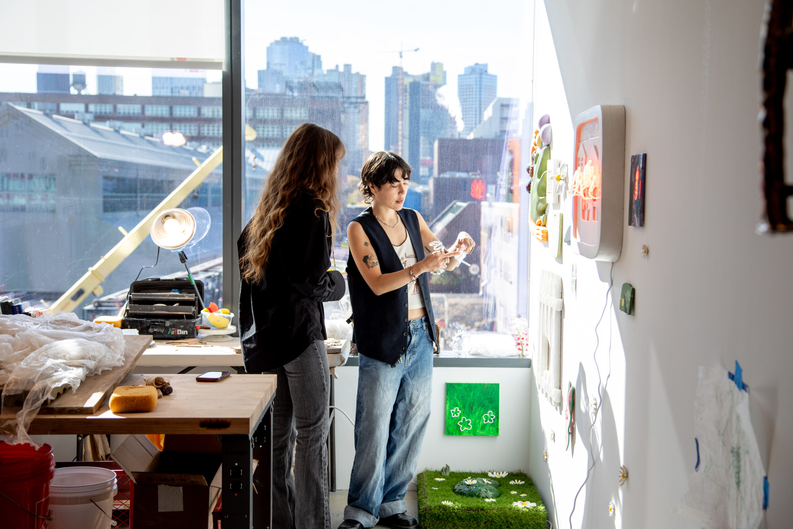 Two people standing in a brightly lit art studio with large windows overlooking an urban skyline. One person with long hair wears a black jacket, while the other, with short hair and tattoos, wears a vest and jeans, examining an object in their hands. The studio is filled with art supplies, a workbench, and wall-mounted artwork, including a patch of artificial grass with small flowers and a green canvas with painted clovers.