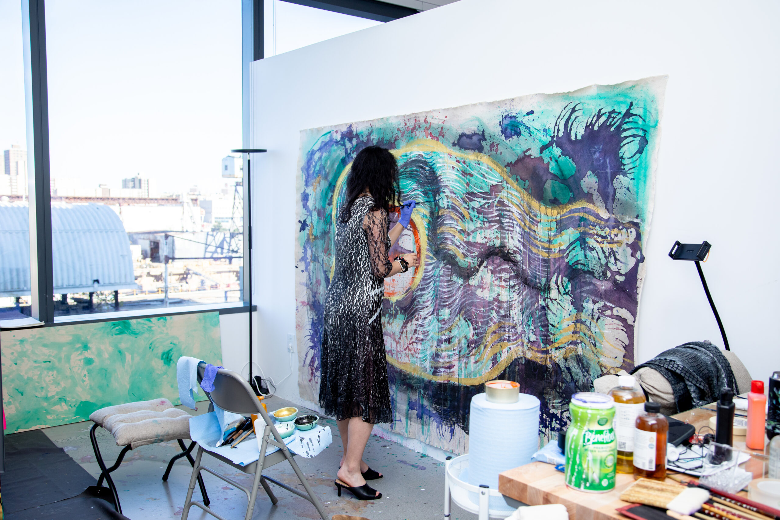 An artist wearing a patterned dress and gloves works on a big, vibrant, abstract painting in a brightly lit studio with large windows. Around the studio, art supplies are scattered on tables and chairs, including brushes, bottles, and containers.