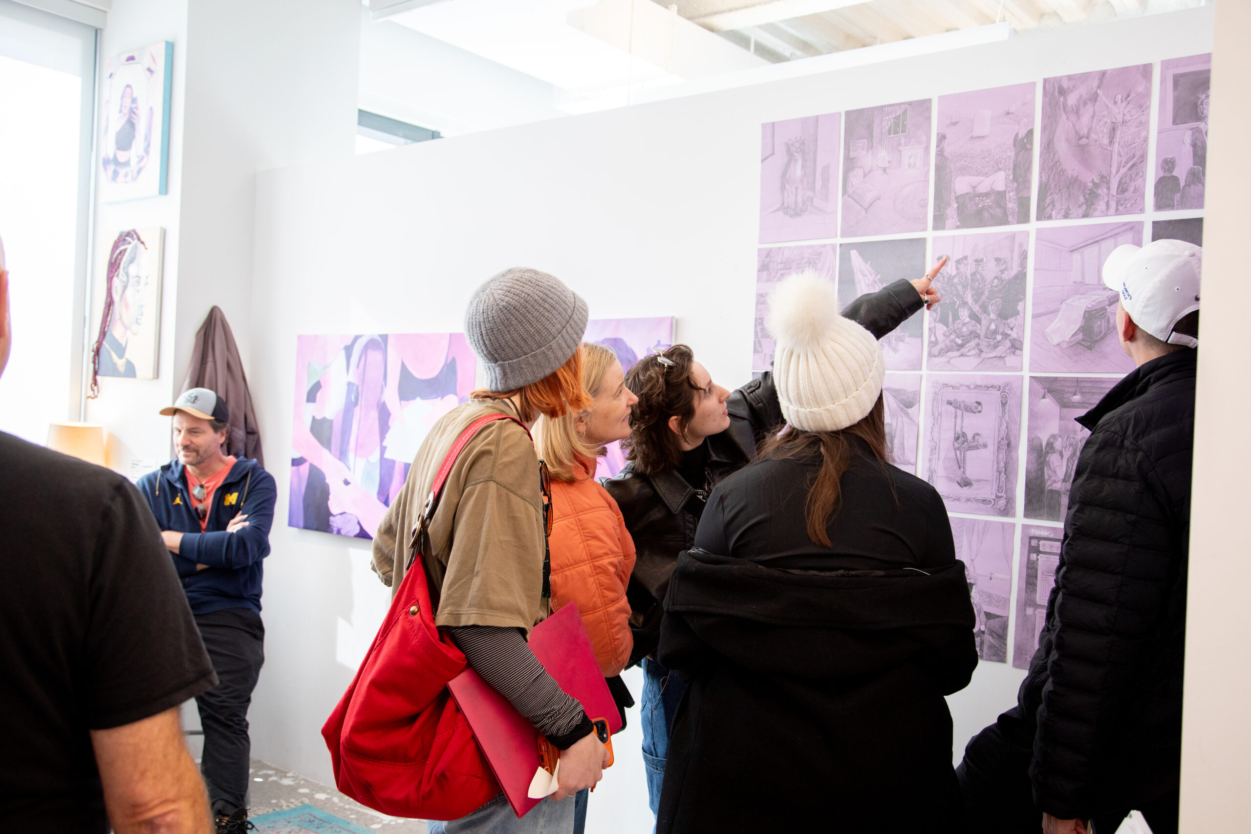 A group of people gathered in a studio, observing a purple-tinted artworks. One person points towards the artwork. The viewers are dressed warmly and one person holds a bright red bag. Additional artwork is visible on adjacent walls.