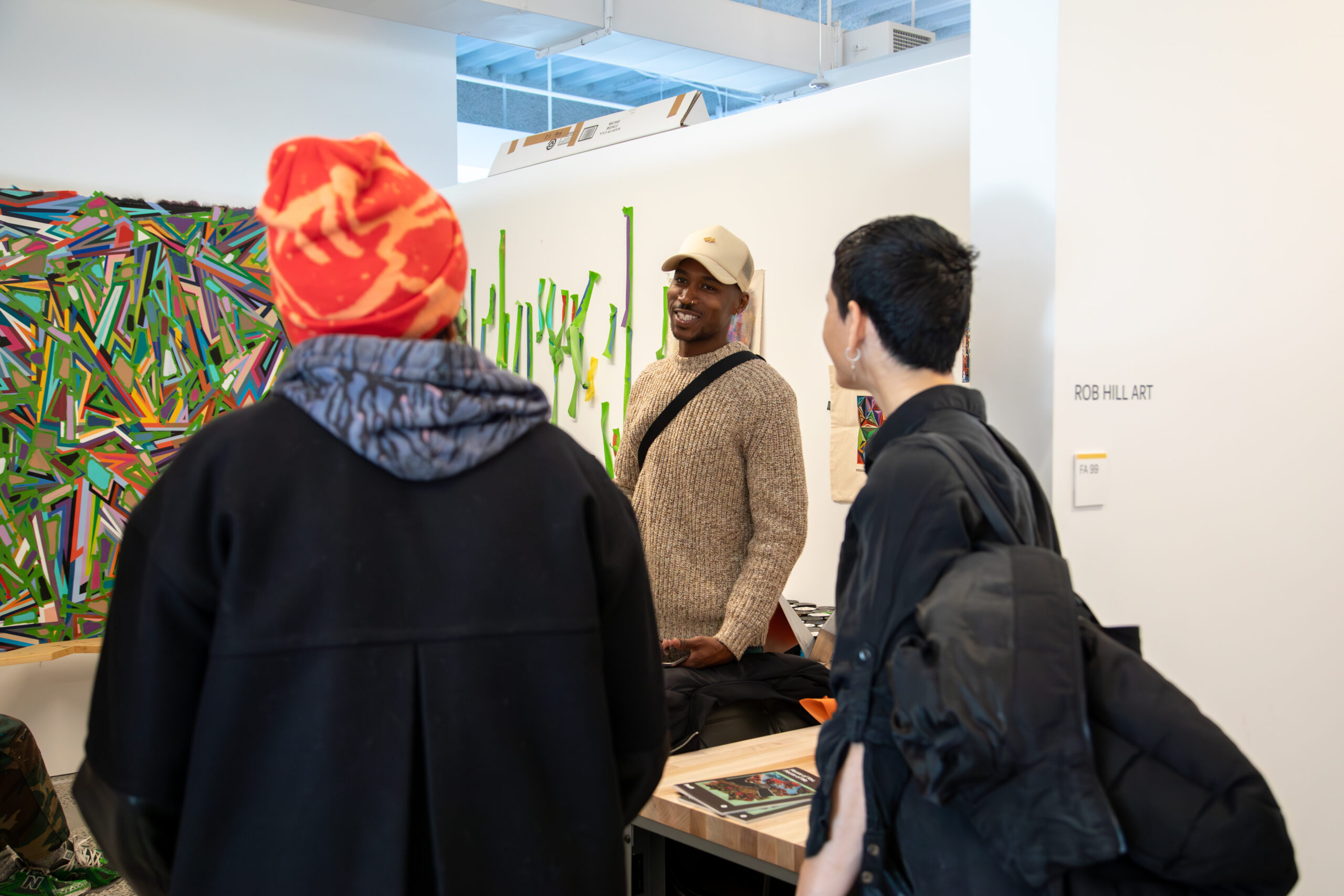 A group of people engaged in conversation at an art studio. One person, wearing a beige hat and a beige sweater, smiles while talking to two others, one in a vibrant orange and red beanie and the other with short dark hair and a black jacket. Colorful, abstract artwork featuring layered lines and green textured shapes is displayed on the walls around them. A small sign on the wall reads 'ROB HILL ART.'
