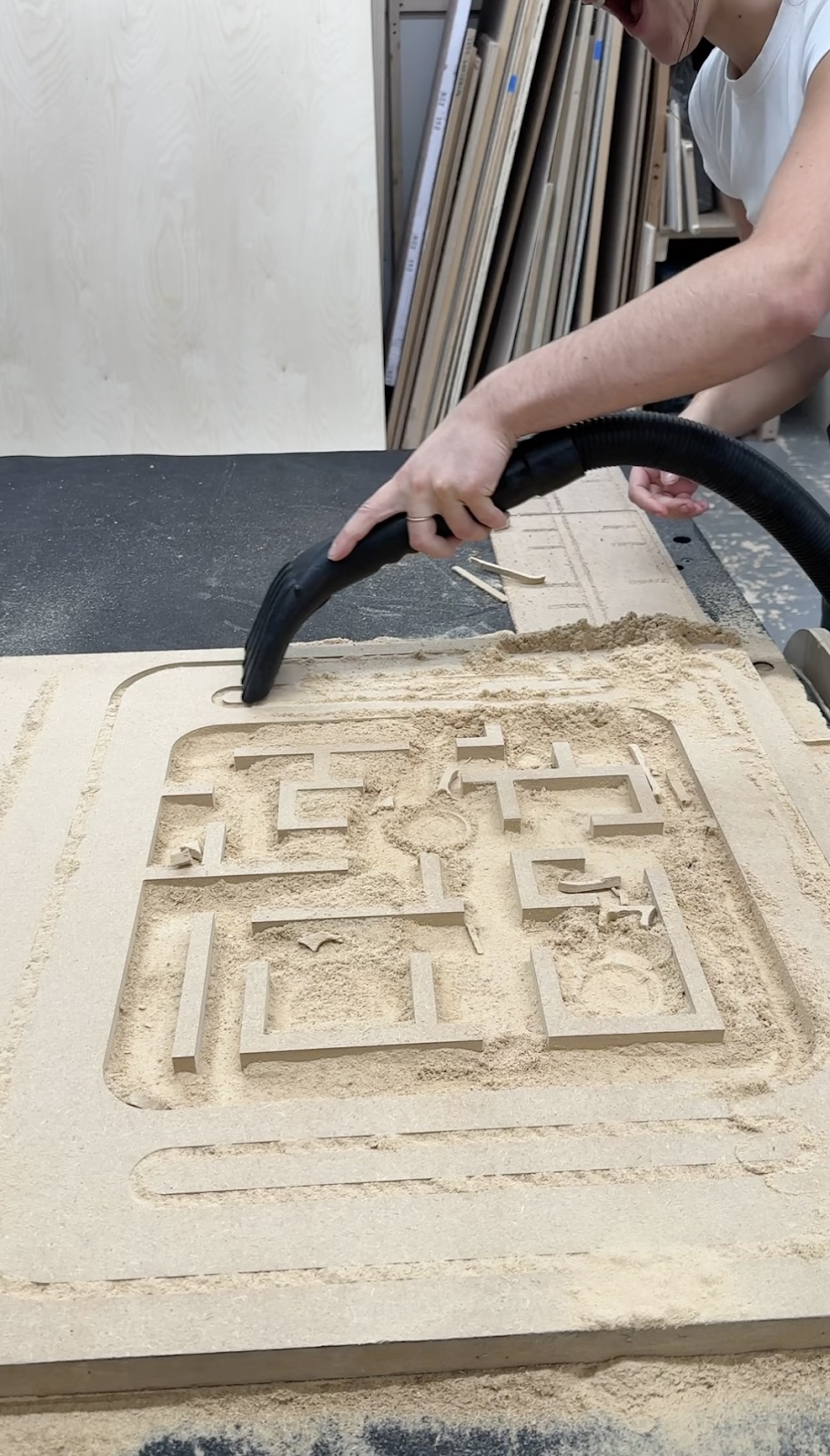 A close-up of a CNC-cut wooden board in a workshop, with a person vacuuming up the sawdust. The board has maze-like pathways and shapes cut into it, with layers of sawdust scattered around the design.