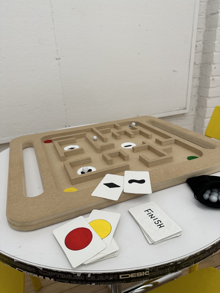 A completed wooden maze board on a round table, featuring colored dots (red, green, yellow, and blue) in the corners. Playing cards with shapes and colors, as well as small white game pieces, are placed nearby, suggesting a color and shape-matching game.