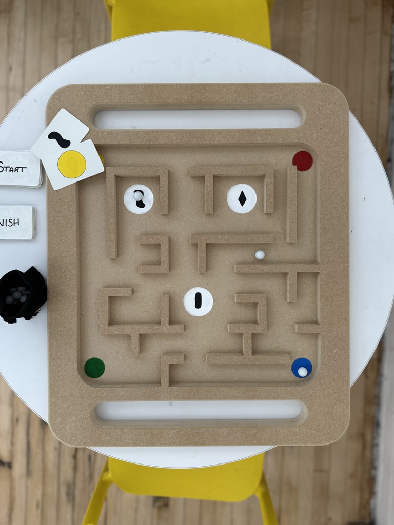 A top-down view of a wooden maze game board on a round table with yellow chairs. The board has intricate pathways with colored dots at the corners and circular spaces for game pieces. A small stack of cards labeled 'START' and 'FINISH' lies next to the board along with additional cards featuring shapes and colors.