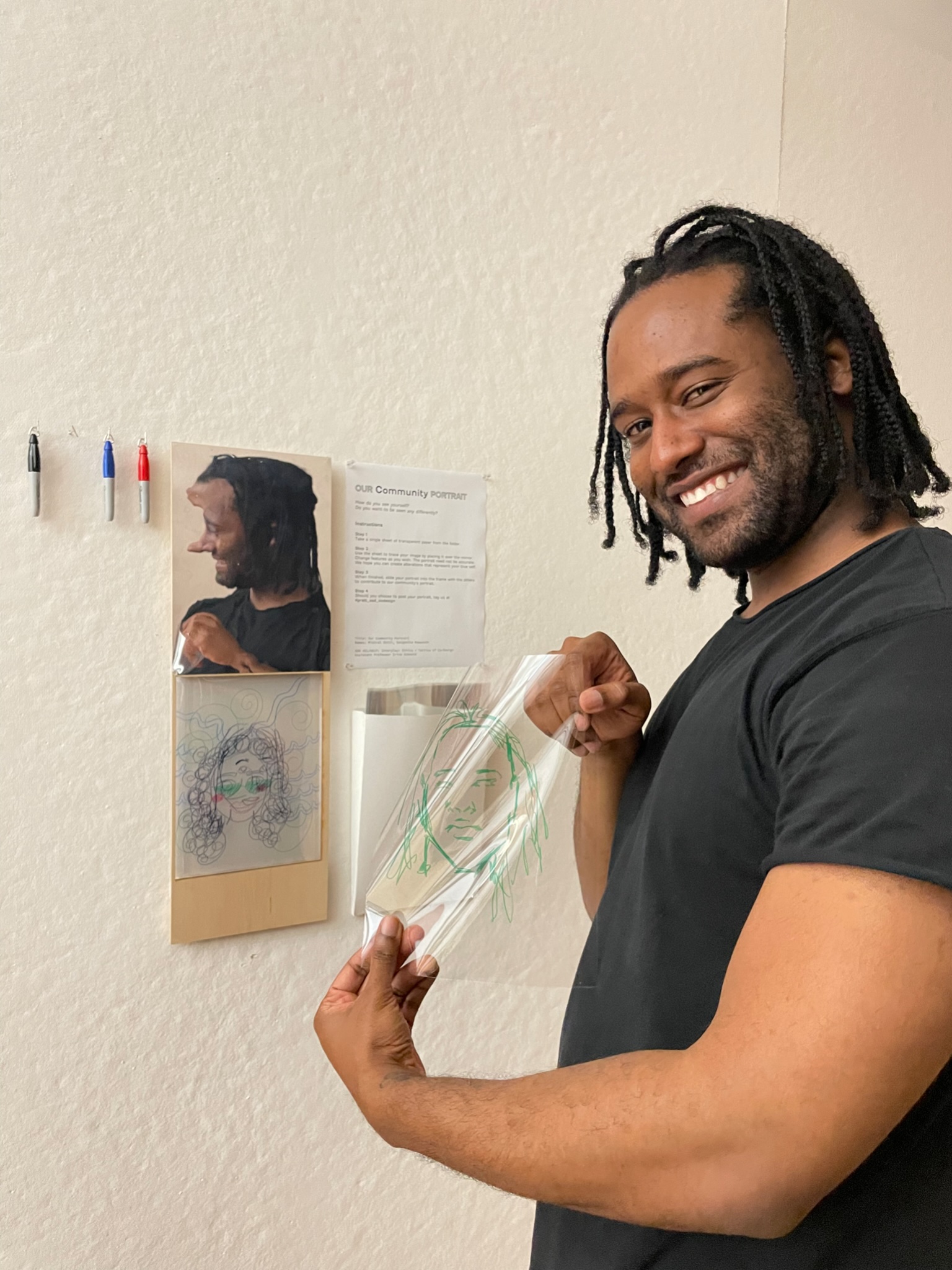 A person smiling and holding a transparent sheet with a green line drawing of a face in front of an interactive art wall labeled 'Our Community Portrait.' Behind them, there are several other line drawings layered on transparent sheets, as well as a photograph and instructions for creating community portraits. Markers are available on the wall, inviting participants to contribute their own drawings to the display.