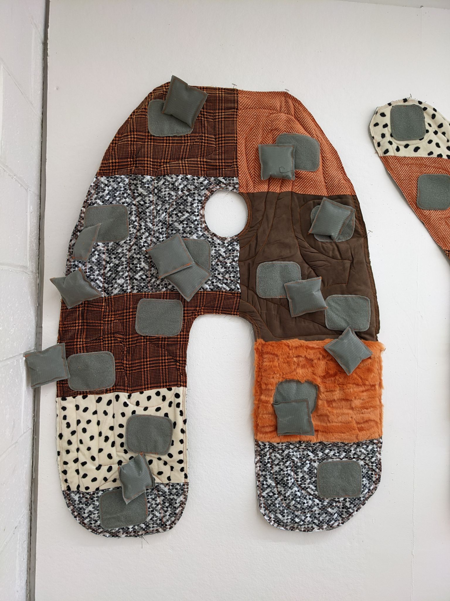 Close-up of a large fabric-covered letter 'A' from the word 'PLAY' in an art installation. The letter is made from a patchwork of different textured fabrics, including patterns in brown, orange, and black-and-white, with multiple small, padded squares attached, adding a tactile element.