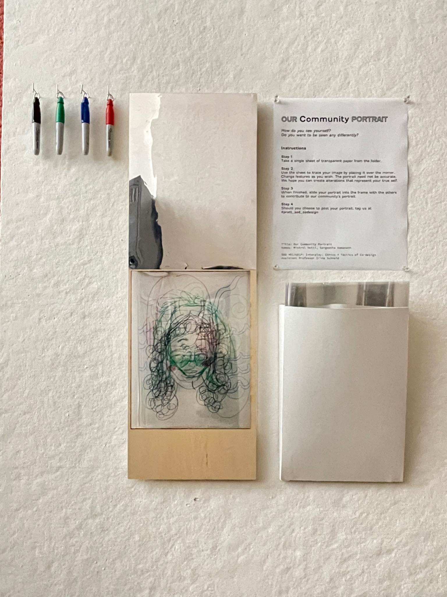 A wall display titled 'Our Community Portrait' featuring instructions for creating a self-portrait using transparent sheets. Four colored markers (black, green, red, and blue) are hanging on hooks to the left. Below the instructions, there is a mirror for participants to trace their reflections onto transparent sheets. Several layered line drawings of faces on transparent sheets are placed in a holder beneath the mirror, showcasing contributions to the community portrait project. A larger pocket on the right holds additional blank sheets for participants.
