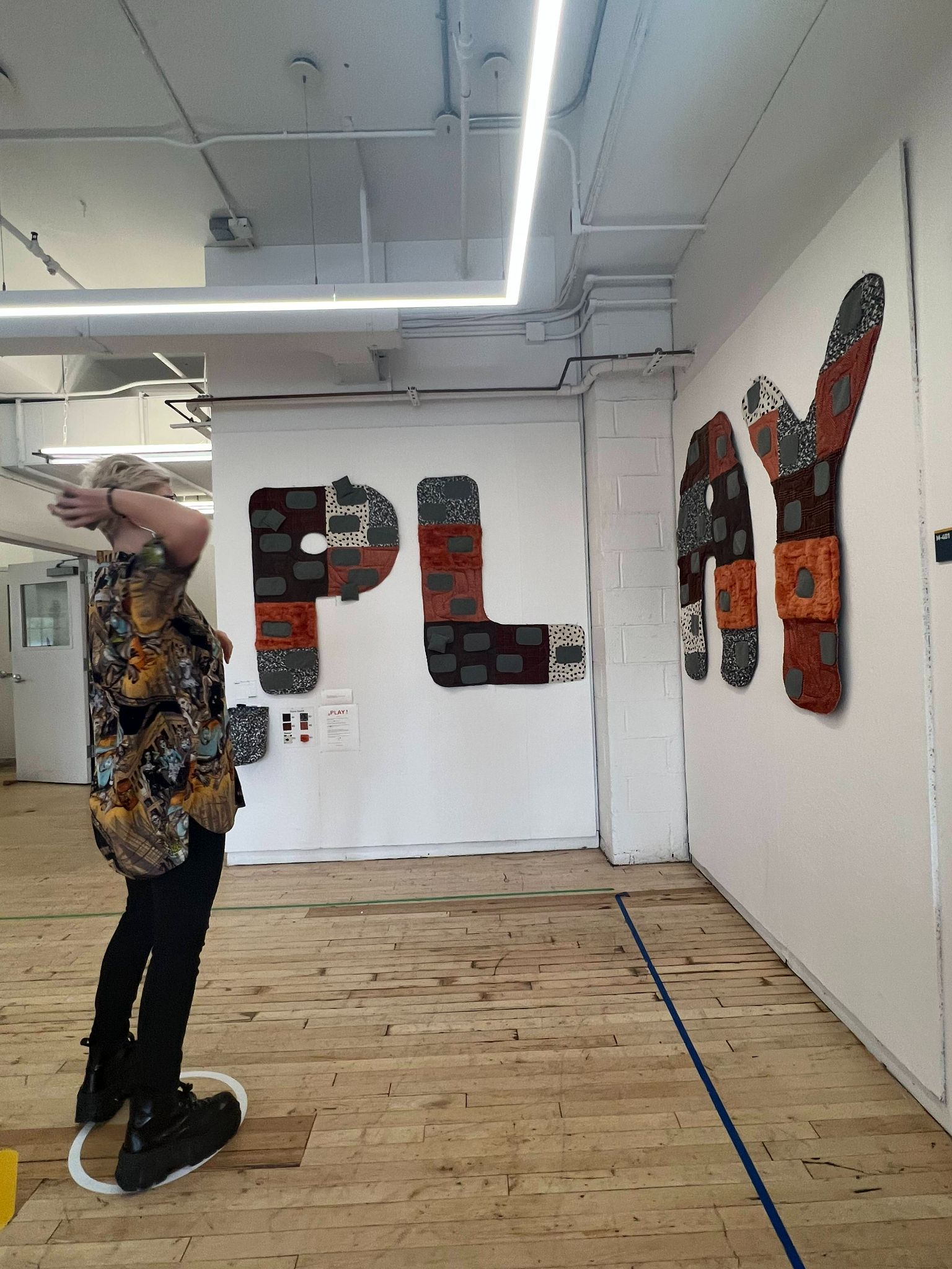 A person standing with their arm raised, looking at a wall installation of large letters spelling 'PLAY.' Each letter is covered with fabric in various textures and earthy tones, with patches and raised shapes giving it a tactile look. The installation has a playful, interactive vibe, inviting engagement.