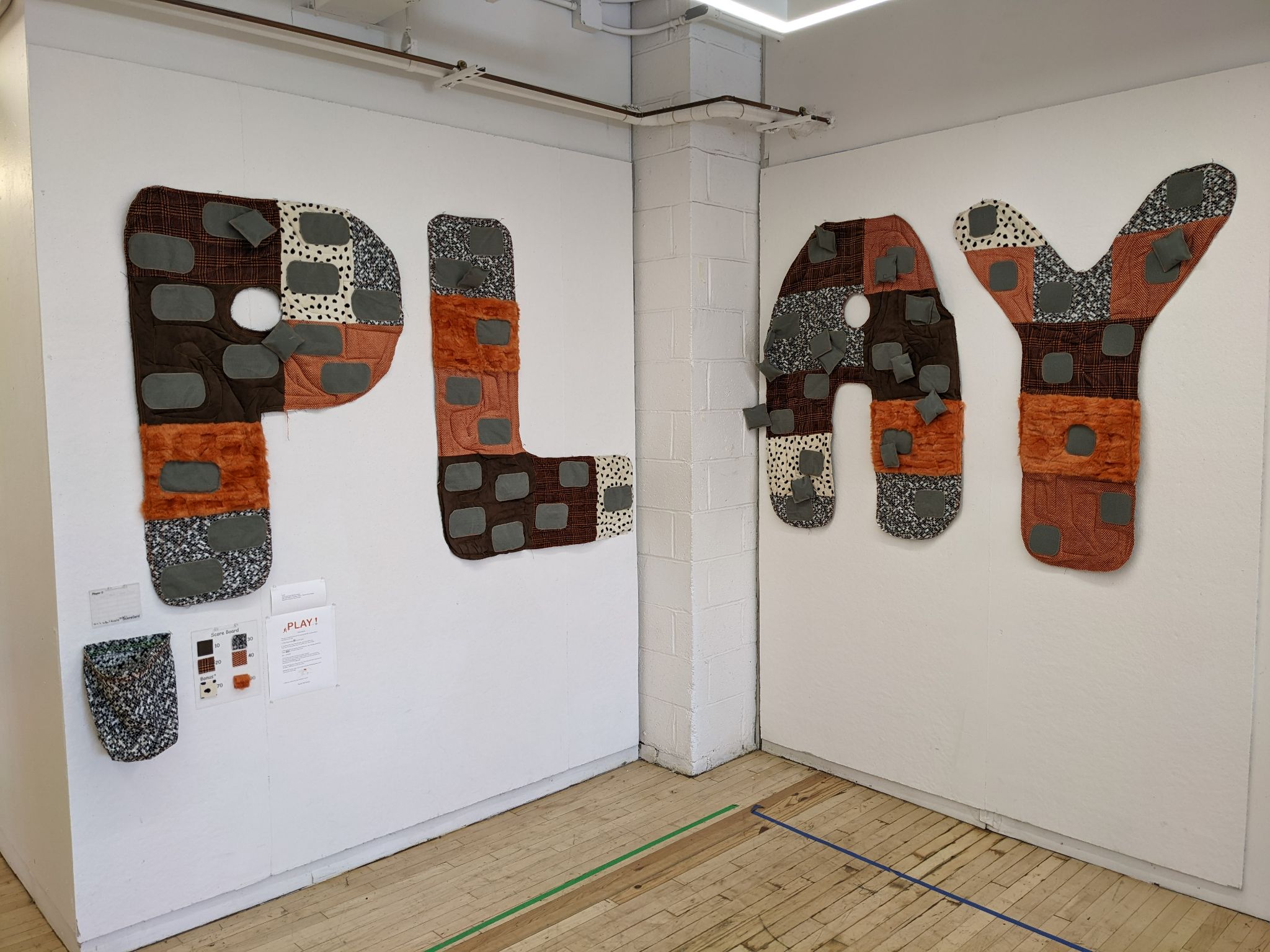 Large, fabric-covered letters spelling out 'PLAY' are mounted on a white wall. Each letter is crafted from different textured fabrics in earthy tones like brown, orange, and gray, with various patchwork patterns and raised square shapes. A small informational sign and sample fabrics are displayed on the wall below the letters.