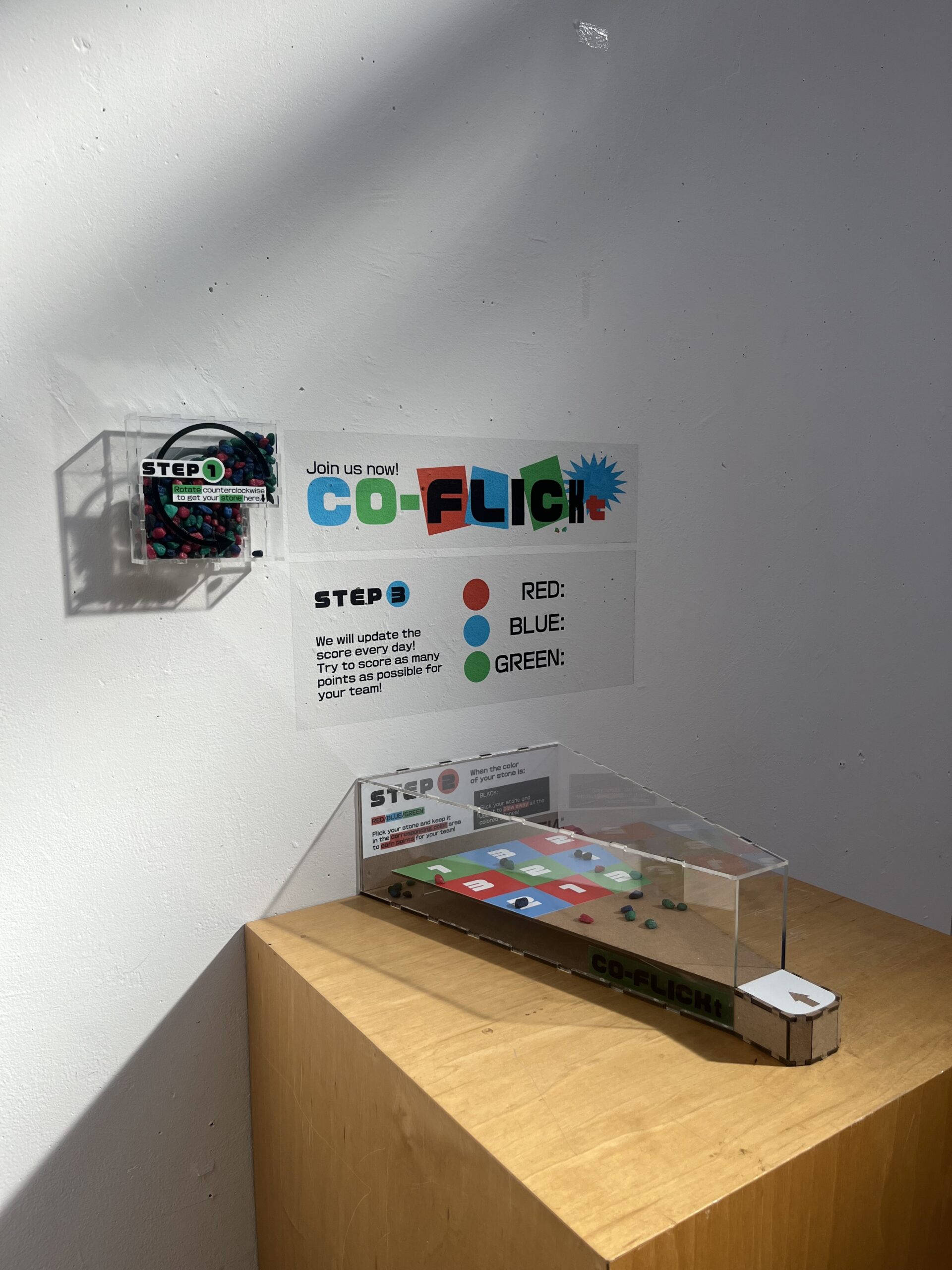 A display setup on a light wooden pedestal featuring an interactive activity labeled 'CO-FLICKt’. A sign on the wall provides instructions with three colored circles (red, blue, and green) for tracking scores, along with 'Step 1' and 'Step 2' instructions. To the left is a small container labeled 'STEP 1' filled with colored counters. The main activity board, encased in a clear cover, has a colorful grid with numbered and colored squares, where participants can flick counters. Shadows cast across the setup add dimension to the display.