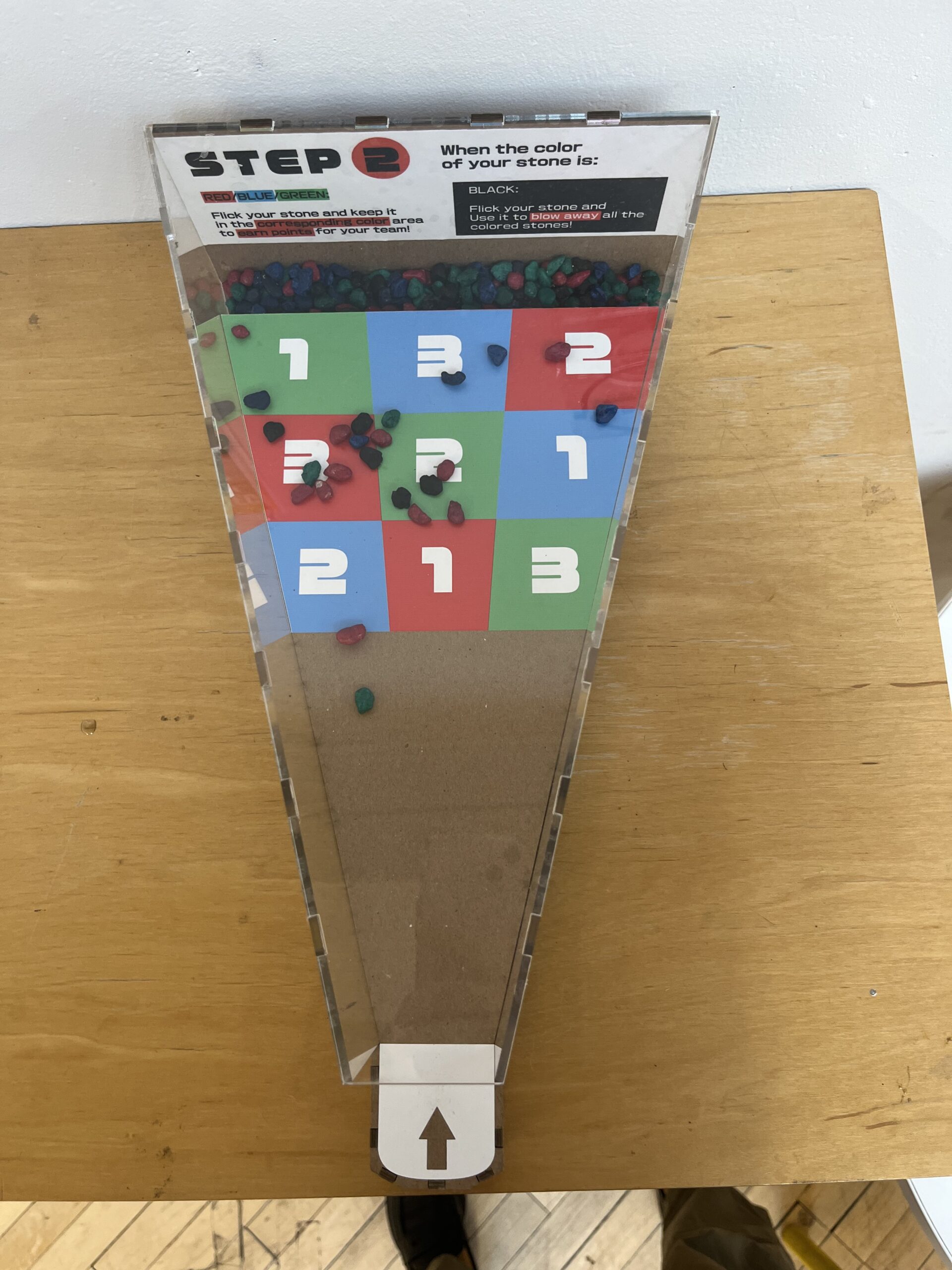 Close-up of an interactive game board titled 'STEP 2' on a wooden surface. The board features a colorful grid of numbered squares in red, blue, and green, each with large white numbers. Small colored stones, in red, blue, and green, are scattered across the grid. Instructions at the top indicate that players should flick their stone and keep it in the same colored area to earn points, with additional instructions for black stones. An arrow at the bottom of the board points toward the grid, suggesting the direction for players to flick their stones.