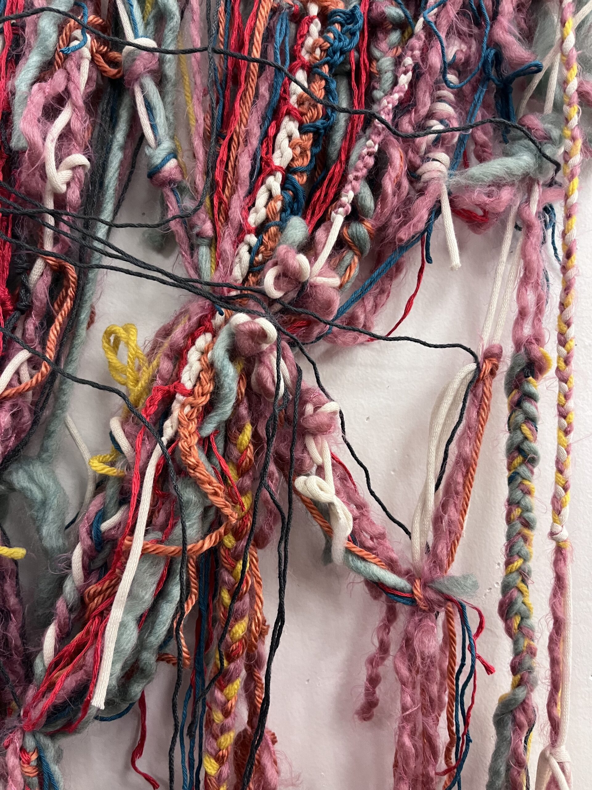 A close-up of a textured art piece made from various intertwined yarns and fibers in shades of pink, blue, red, yellow, and white. The fibers are woven, knotted, and twisted together, creating a complex, layered, and tactile appearance.