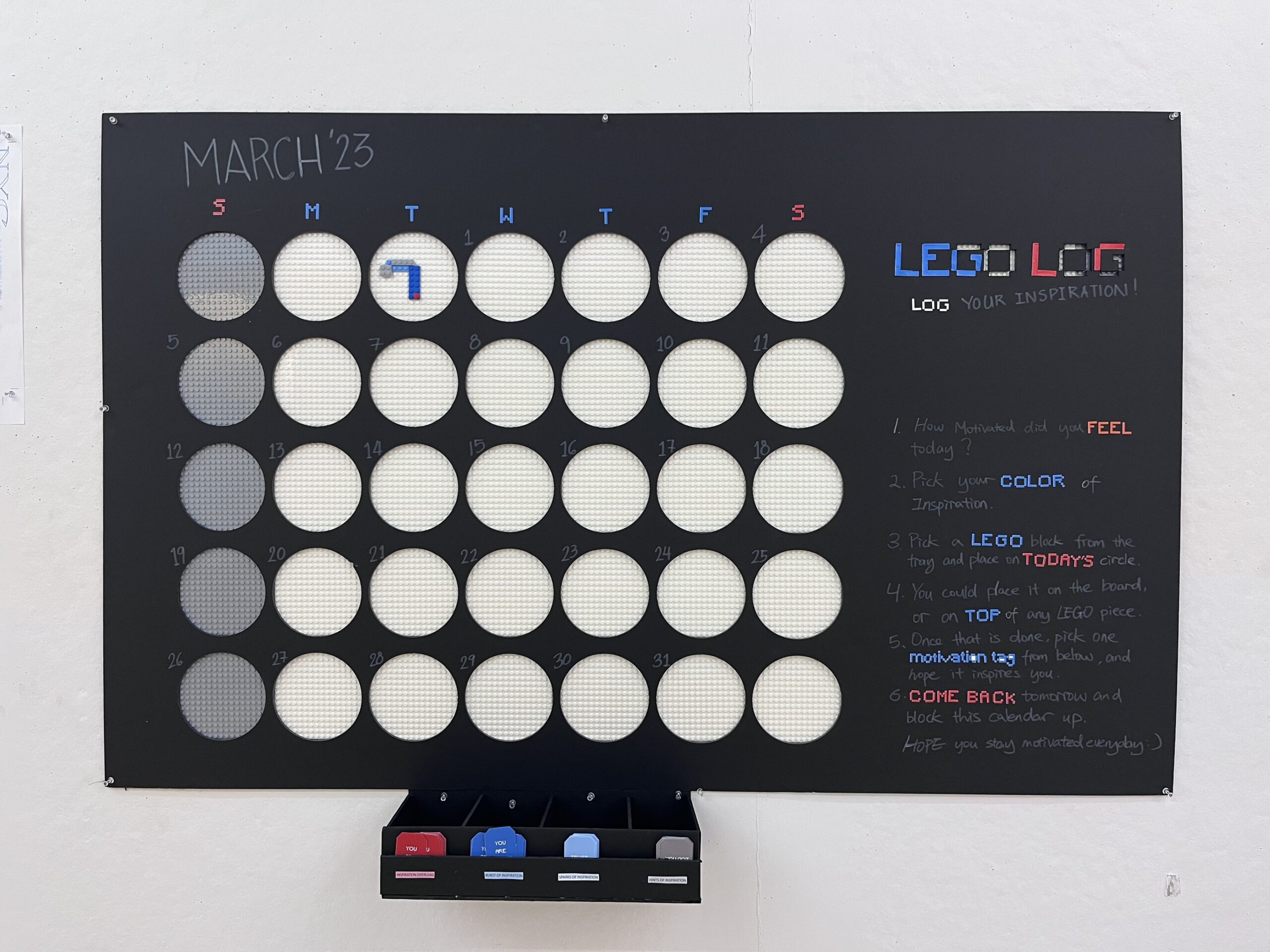 A wall-mounted interactive calendar titled 'LEGO LOG' for March 2023. Each day of the month is represented by a circular pegboard space, some of which have LEGO pieces attached. Instructions on the right side explain how to log daily motivation by selecting a color of LEGO and placing it on the current day's circle. Below the calendar, there’s a tray holding red, blue, and gray LEGO pieces, labeled with motivational prompts. The activity encourages participants to return each day and track their inspiration visually using LEGO bricks.