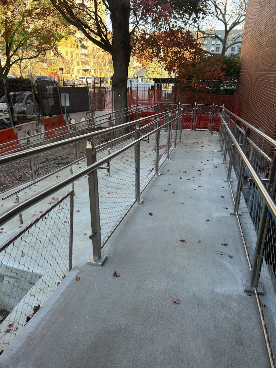 Photo of a concrete wheelchair-accessible ramp with stainless steel handrails and chain-link fencing. The ramp leads down to a gated area, surrounded by construction barriers and trees with autumn-colored leaves.