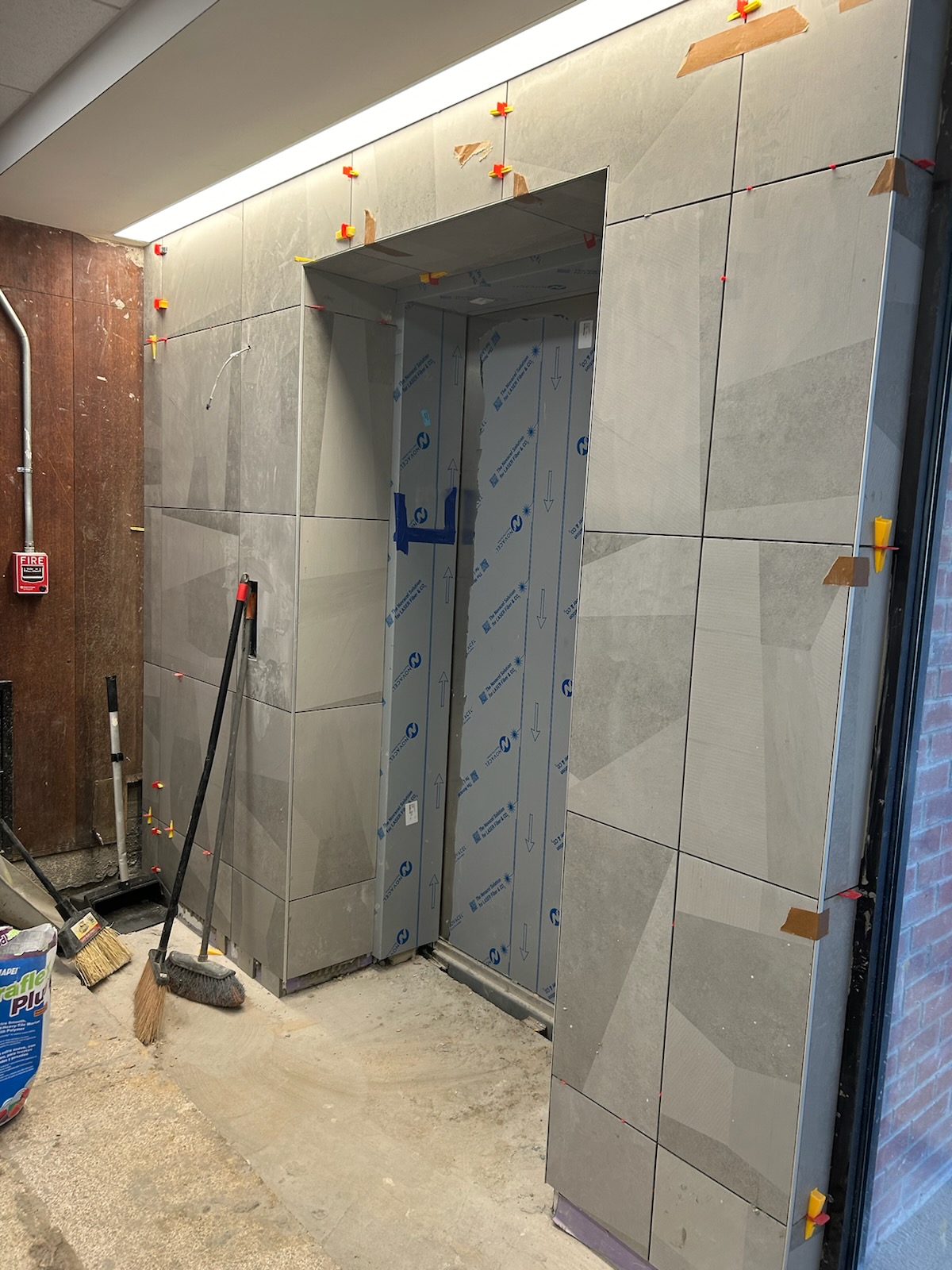 Construction site of an elevator installation surrounded by large gray tiles with visible adhesive spacers. Tools, such as a broom and dustpan, lean against the partially finished wall.