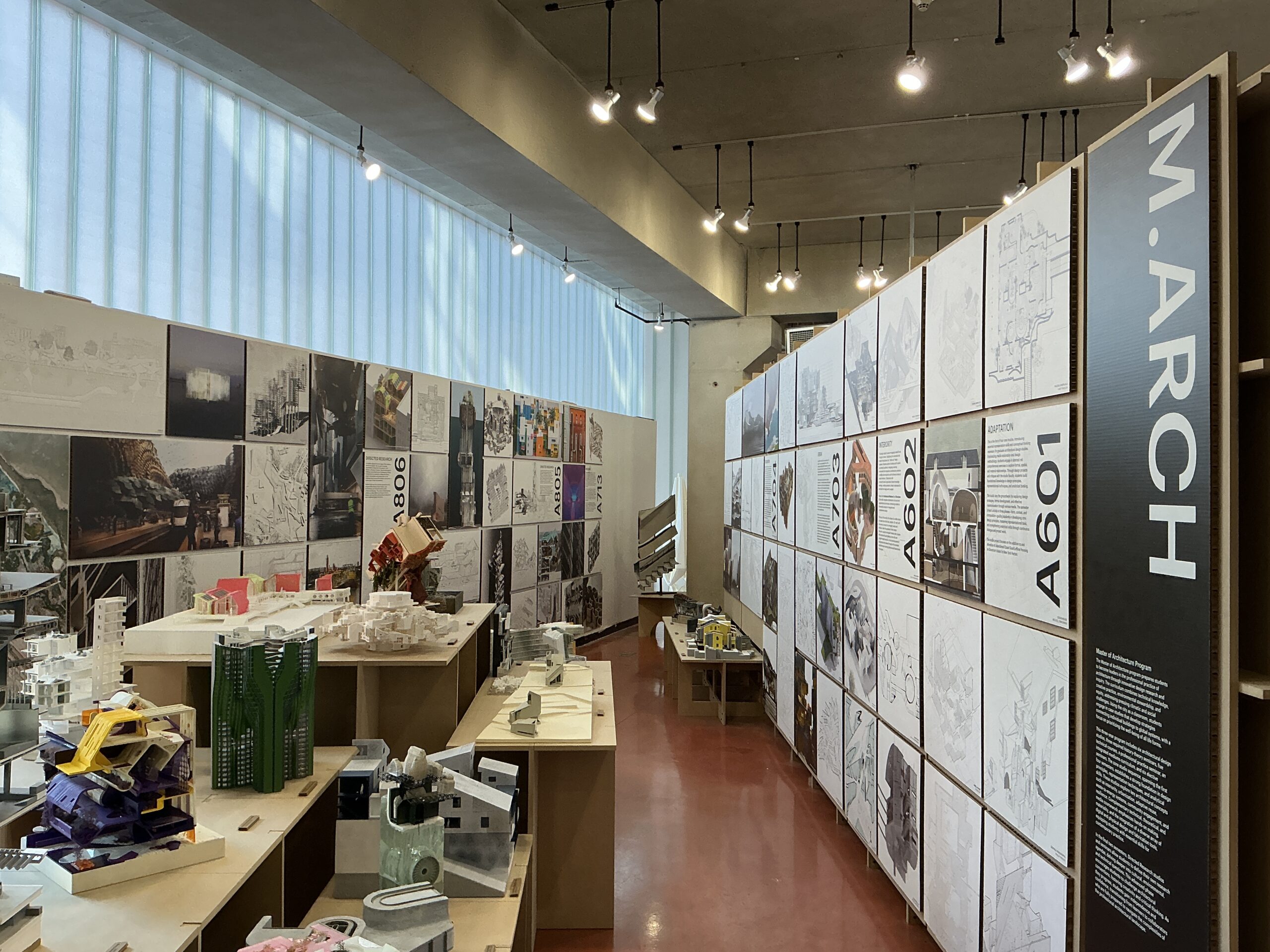 An architectural exhibition room featuring wooden tables with intricate physical models of buildings and structures. The walls are covered in large presentation boards displaying architectural plans, sections, and colorful renderings. The room has red flooring, brick and white walls, and is illuminated by overhead lighting. Labels such as 