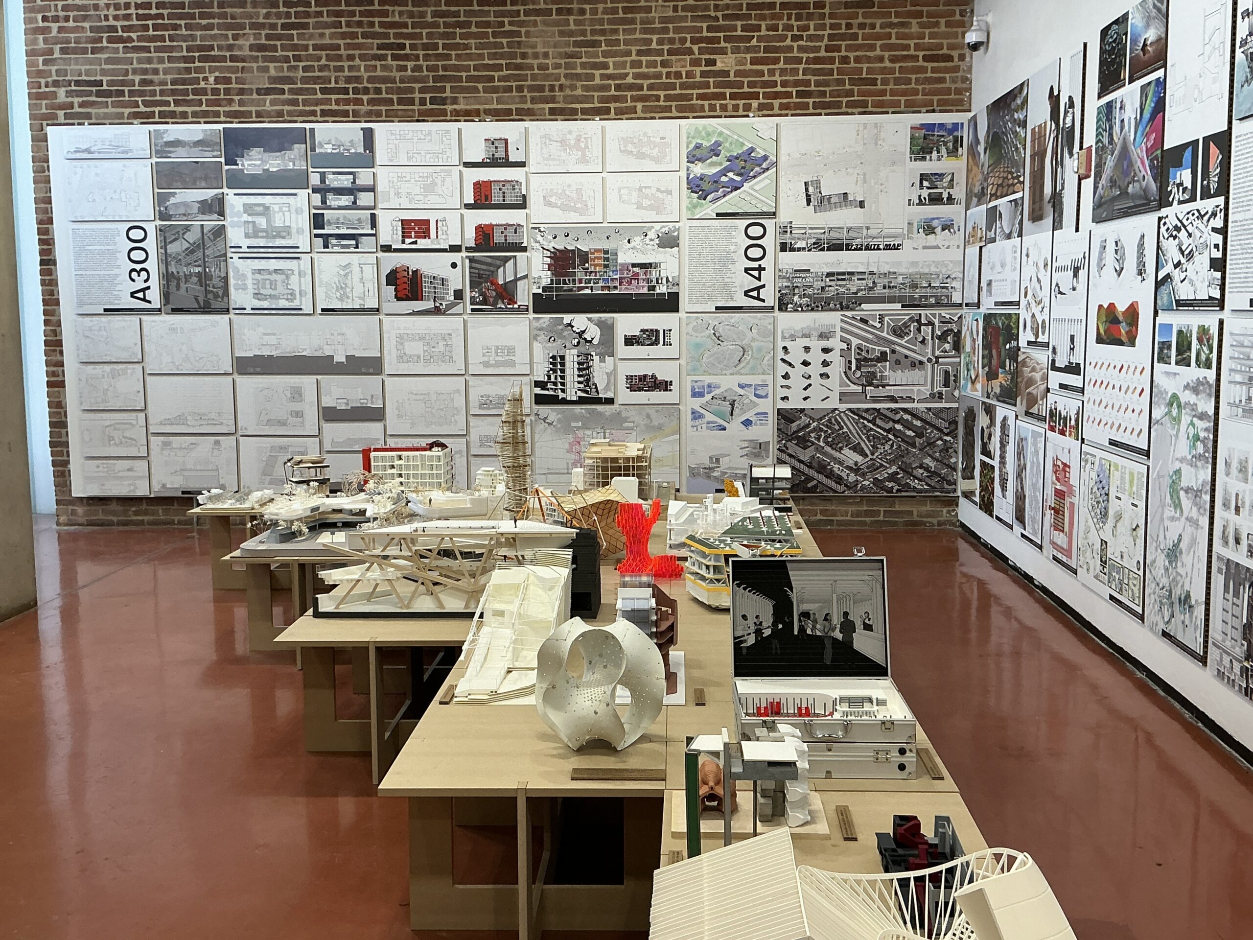 Close-up of several architectural models displayed on wooden tables. The models vary in size and complexity, including sectional building cutaways, high-rise structures, and abstract design interpretations. Behind the models, large boards display detailed architectural plans, perspectives, and conceptual illustrations.