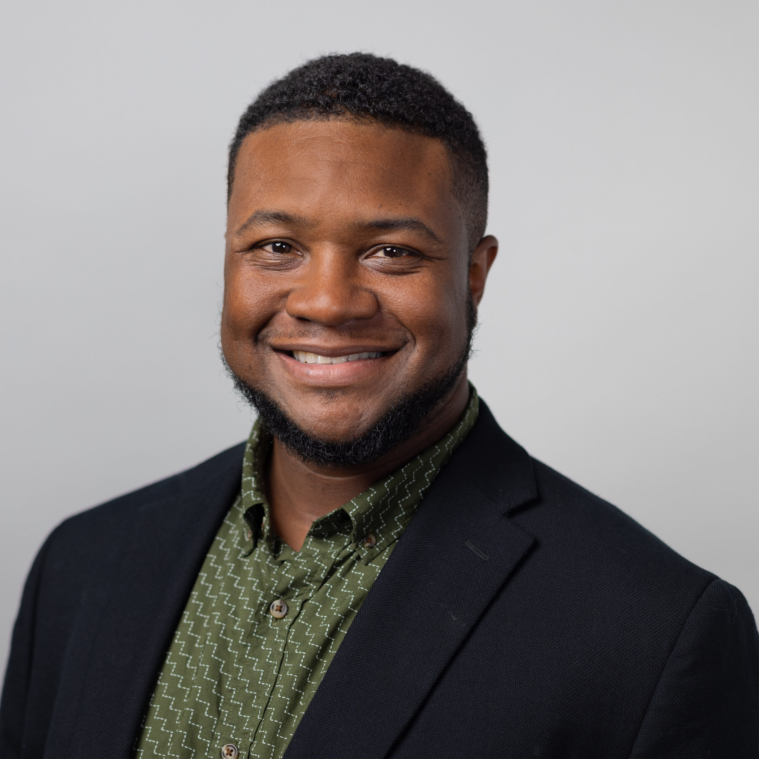 A man with short curly hair and a neatly trimmed beard smiles warmly at the camera. He is wearing a dark blazer over a green patterned button-up shirt. The background is a smooth light gray, giving the portrait a clean, professional appearance.