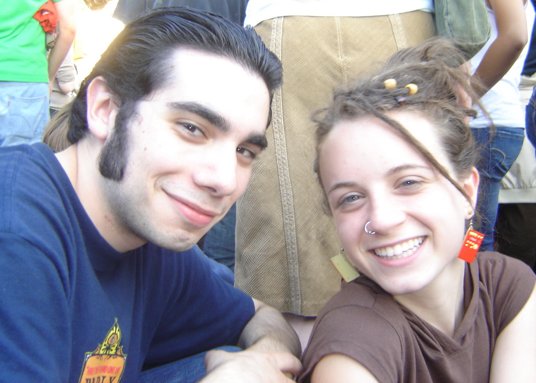 A young man and woman smile at the camera at an outdoor event. The man has dark slicked-back hair and wears a navy blue T-shirt, while the woman has curly hair with small beads woven in, a nose ring, and bright red and yellow earrings. She leans slightly toward him, showing a wide smile. There are people standing behind them.