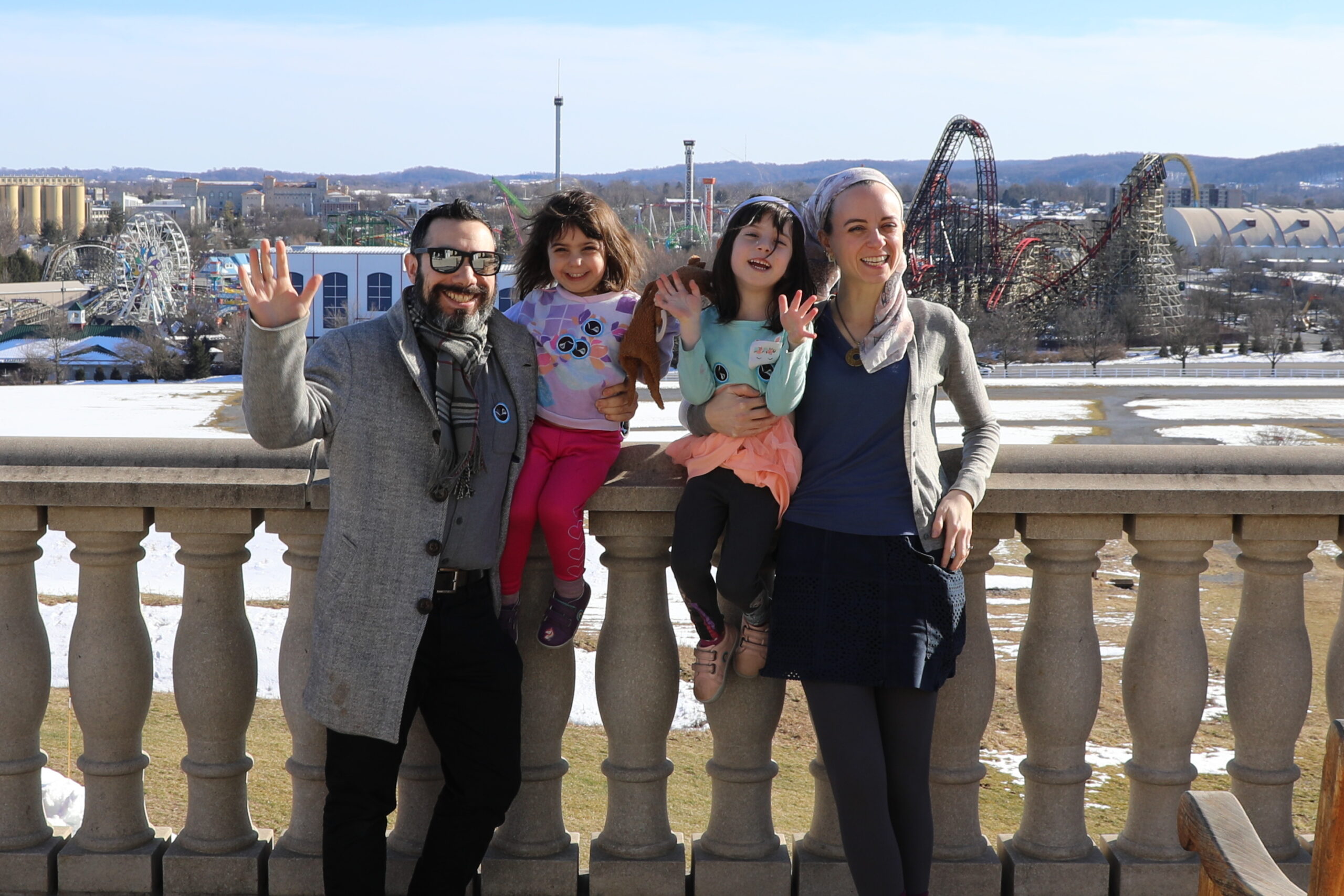 A family of four poses on a scenic overlook with a theme park in the background. The father, wearing sunglasses and a gray coat, waves at the camera. The mother, in a blue top and gray cardigan, holds one of their young daughters, who wears a tutu and raises her hand. Their other daughter, in a colorful shirt, stands between them, smiling. The roller coasters and ferris wheel of the theme park are visible in the distance.