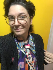 A selfie-style portrait of a person wearing glasses and a vibrant, patterned outfit with a colorful tie. They are smiling slightly and standing in front of a bright yellow wall, exuding a playful and artistic vibe.