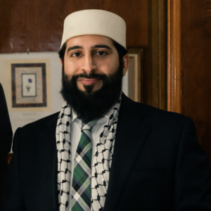A portrait of a person dressed in formal attire, wearing a white hat and a checkered scarf over a suit and tie. They are smiling softly and standing in a warmly lit room with framed artwork visible in the background, creating a professional and welcoming atmosphere.