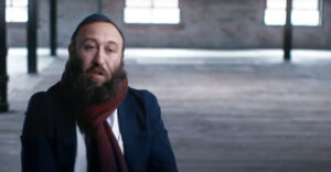 A portrait of a person with a beard and wearing a dark blue suit and a red scarf. They are seated in a spacious, industrial-style room with soft natural light streaming through windows in the background, creating a warm and reflective atmosphere.