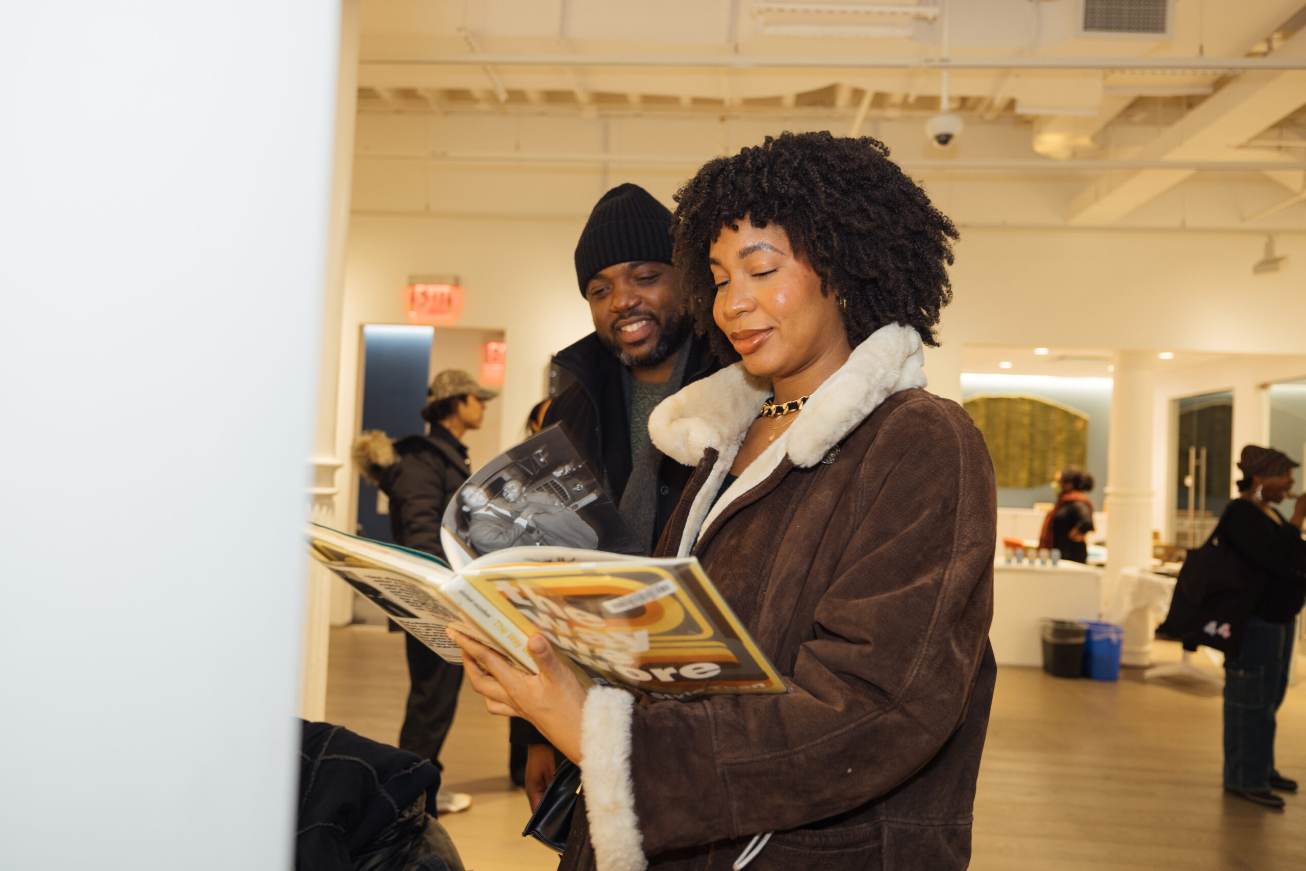 A woman in a brown shearling jacket smiles as she flips through a fashion book, while a man in a black beanie and coat stands beside her, observing the pages with interest.