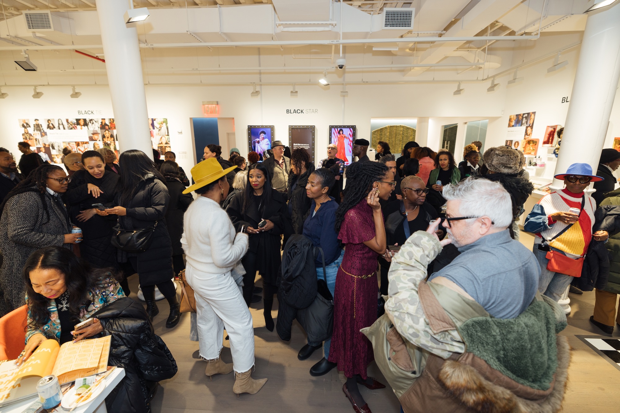 A lively crowd fills an art gallery space, engaging in conversations and admiring fashion exhibits. A person in a mustard-yellow hat and white outfit stands in the foreground.