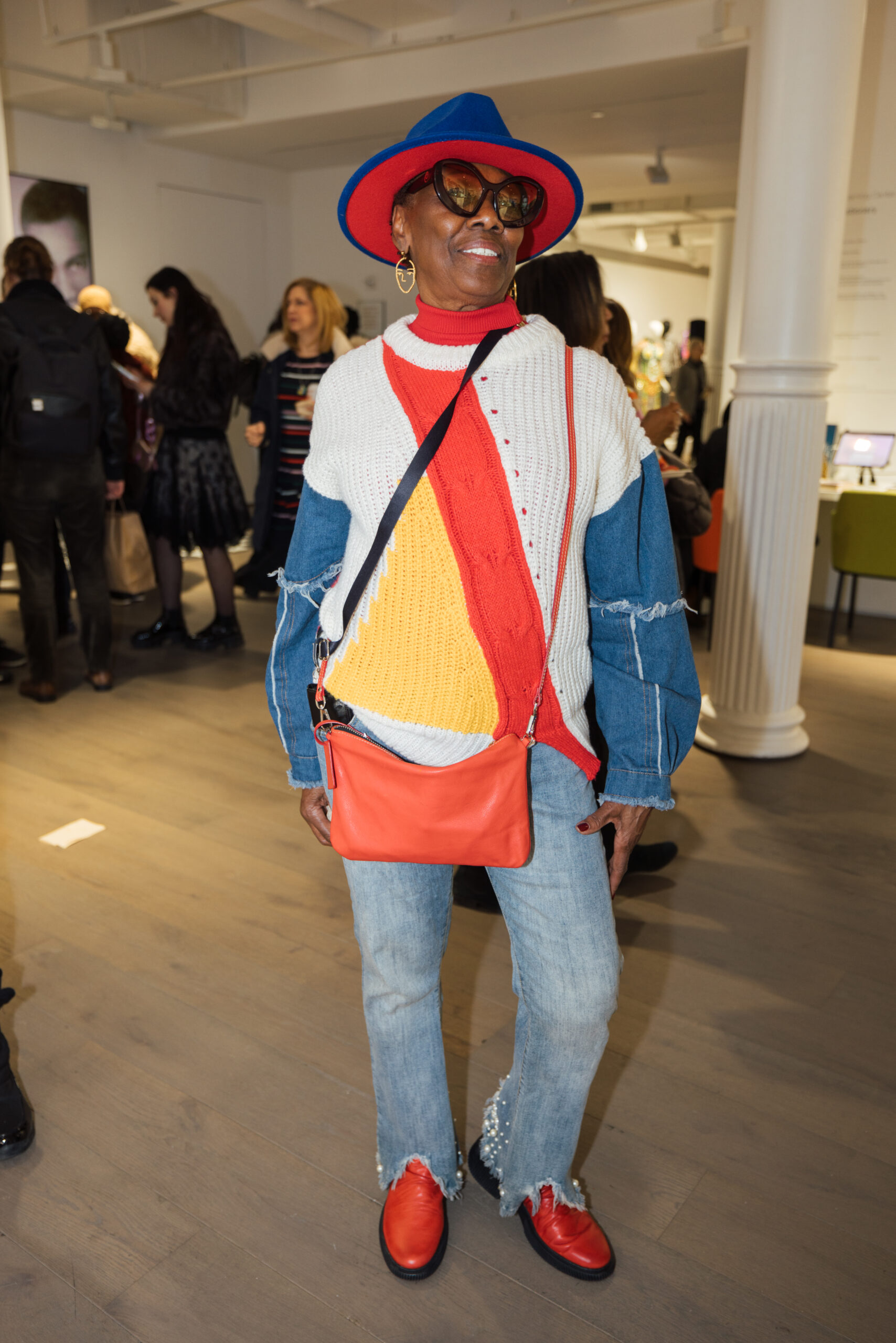 A stylish person poses confidently in a color-blocked knit sweater with red, blue, and white panels, paired with frayed denim jeans, bright red shoes, and a bold red and blue hat, accessorized with a red crossbody bag.