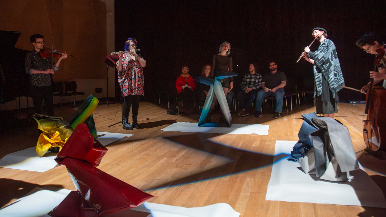 Five people standing in a half-circle performing music—one singing, the others playing instruments—in a dim performance space behind four sculptures arranged in a square. A row of onlookers are seated in the background.