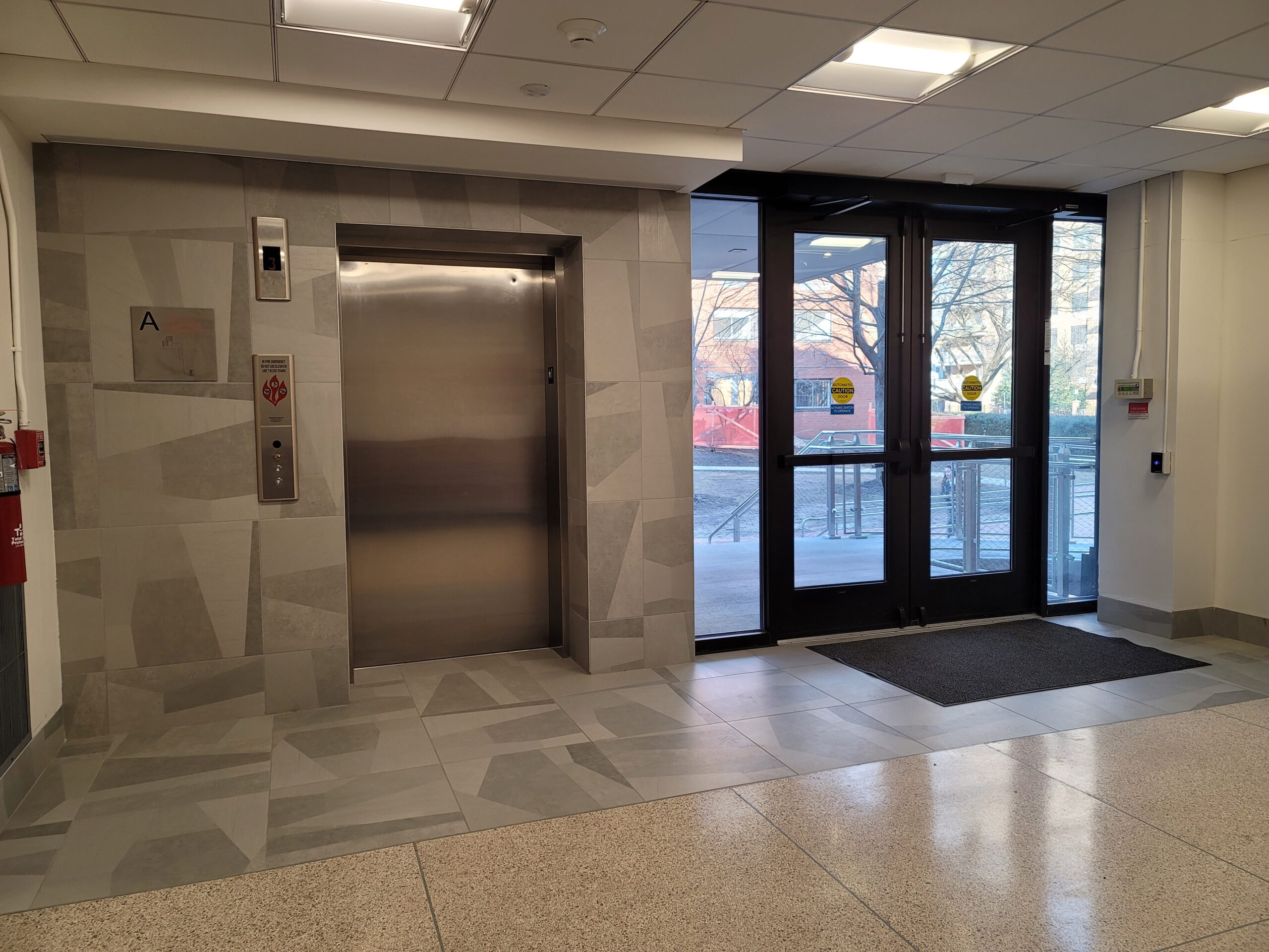 A well-lit indoor space featuring an elevator lobby with a modern design. The walls have a geometric-patterned tile finish, and the floor is a mix of polished stone and tile. A set of double glass doors leads outside, revealing part of a campus walkway. The elevator panel includes accessible signage, and the area is clean and well-maintained.