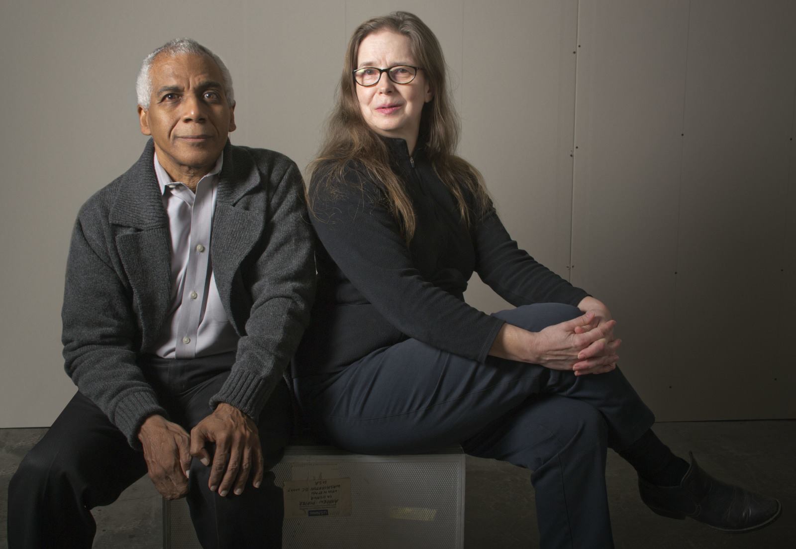 Portrait of Everado Jefferson, a man wearing a gray cardigan and black pants, next to Sara Caples, a woman with red glasses, who also wears dark-colored clothing. They both look at the camera, smiling softly.