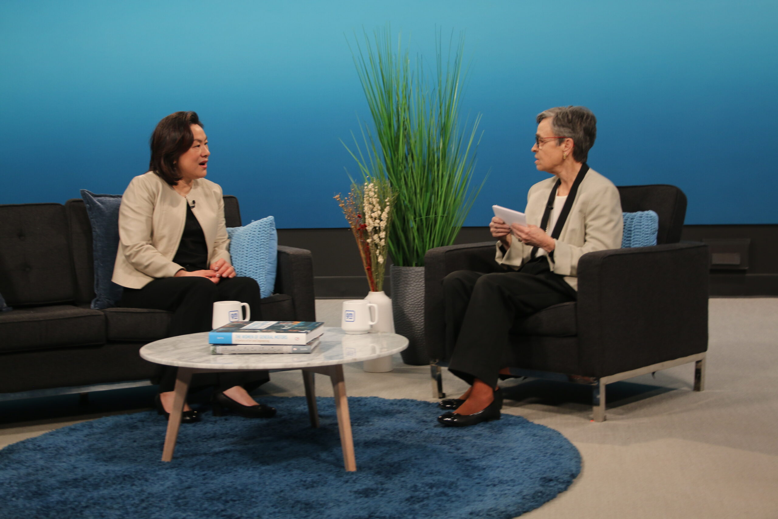 Two women wearing similar outfits—light brown tops and black pants—sit across from each other on a black sofa and chair on a television talk show set. The set backdrop is a blue gradient.
