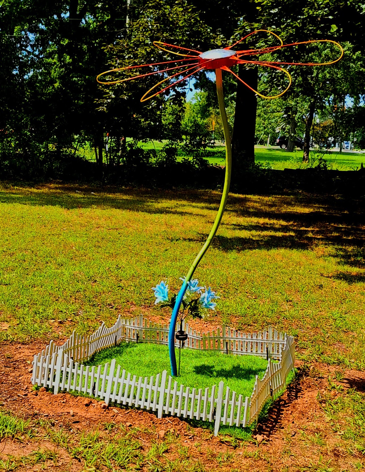 Sculpture of a tall metal daisy with a blue and green stem and yellow-red petals, which stands in a miniature rectangular patch of bright green grass, encircled by a small white picket fence