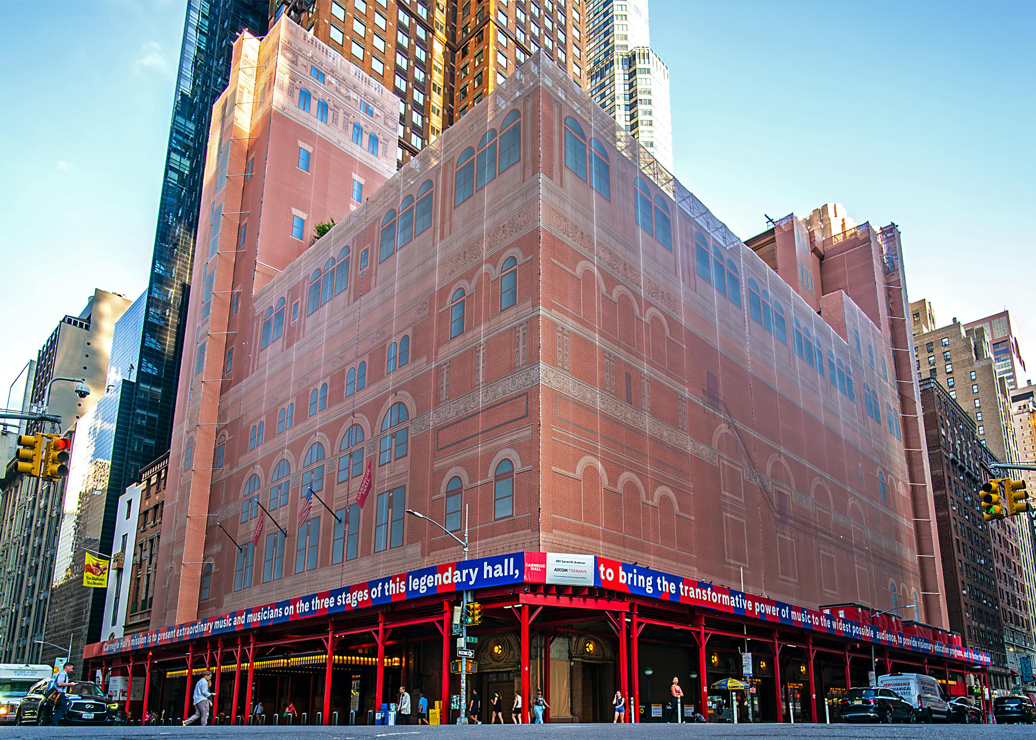 Wide-angle view of Carnegie Hall’s exterior under construction, which has red scaffolding and is obscured by an enclosure that depicts a mock-up of the finished building.