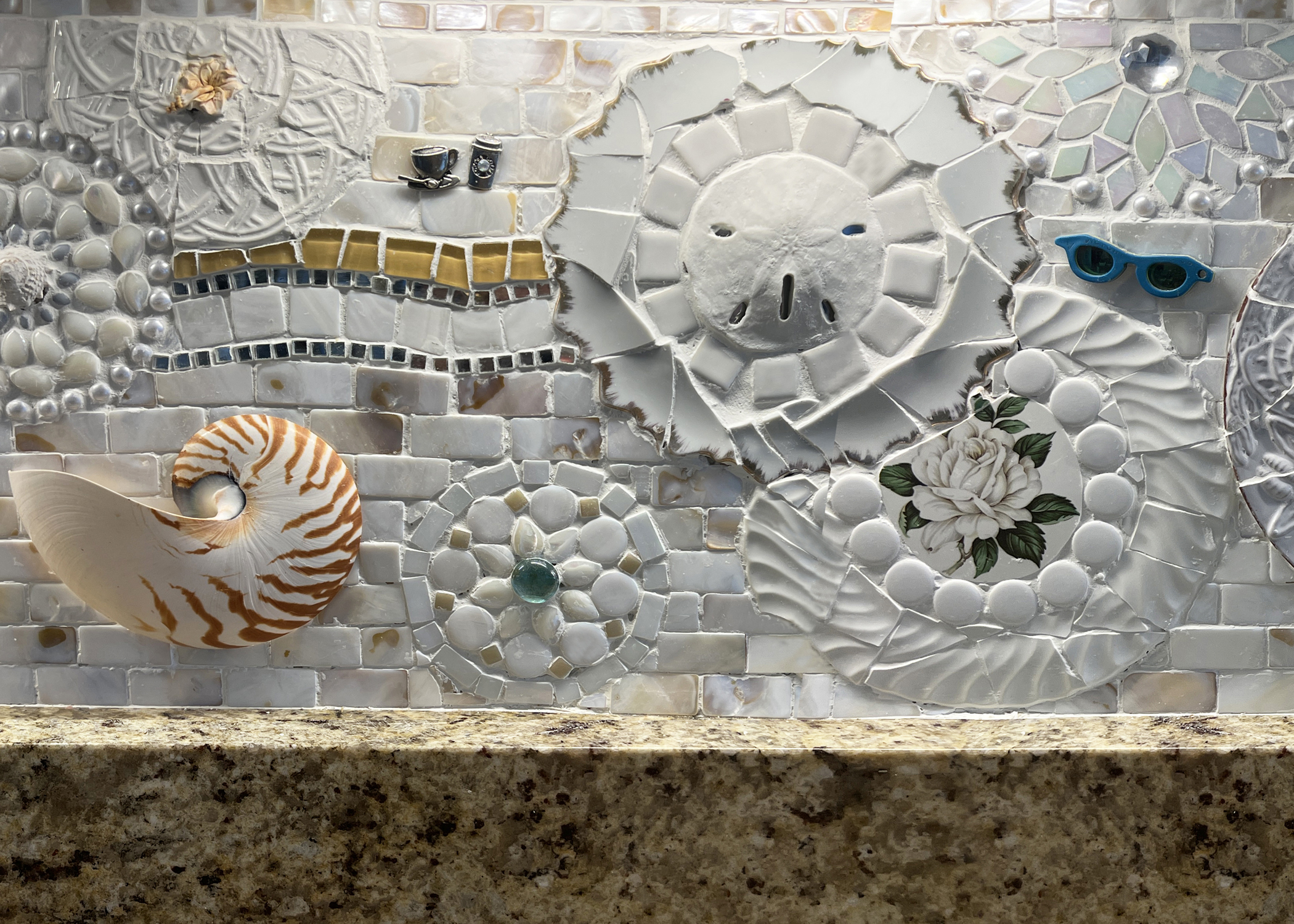 Close-up of a mostly white-colored mosaic kitchen backsplash, which includes collaged tiles, seashells, glass, assorted decorative silver objects, and a teacup adhered to the wall.