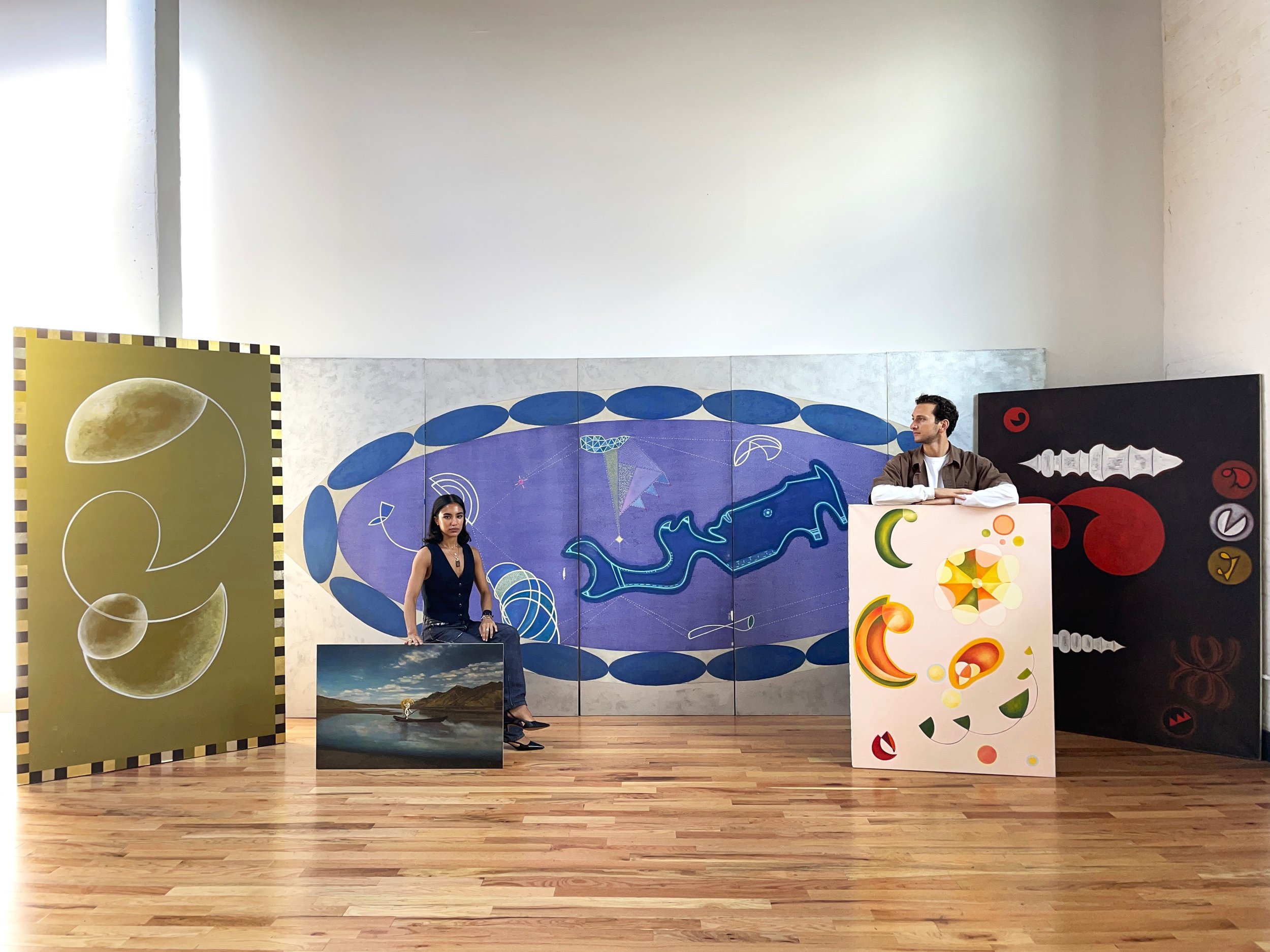 Two people stand behind large painted canvases in a white room, and behind them three more bright, abstract artworks span the length of the wall