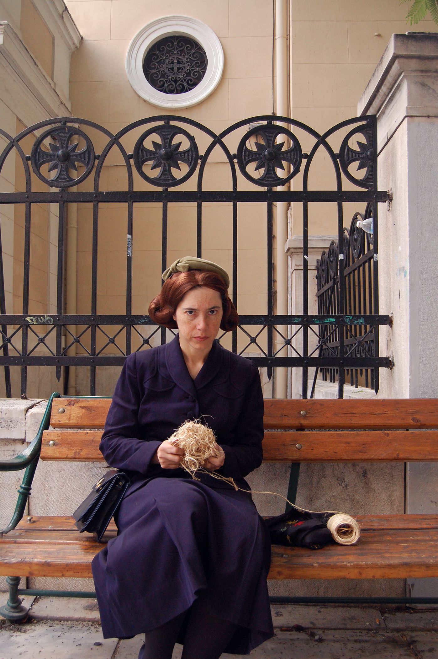 Portrait of a woman wearing an indigo collared dress staring at the camera with a serious expression; she sits on a bench outside of a church fence, holding a knot of yarn