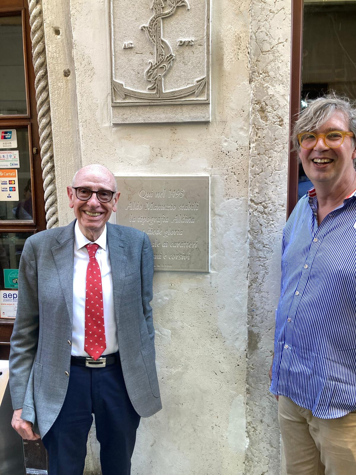Two men smiling at the camera, standing in front of a stone façade with two engraved plaques arranged vertically