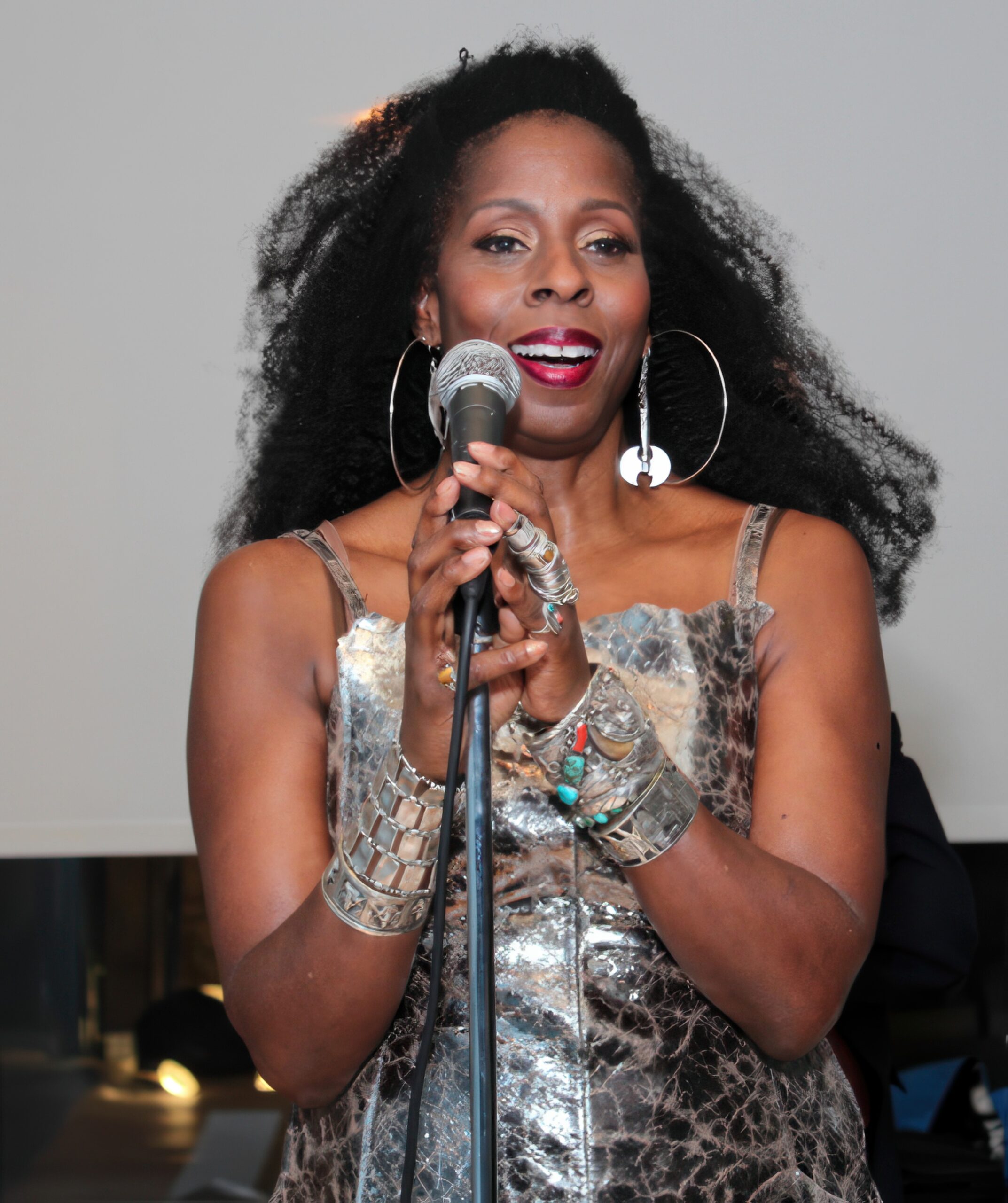 A woman with voluminous hair speaks into a microphone, wearing a metallic silver dress with thin straps. She is accessorized with large hoop earrings, multiple statement rings, and an array of bold metallic and beaded bracelets. Her expression is radiant and confident, with deep red lipstick and a poised stance. The background is softly lit, suggesting an elegant event or performance setting.