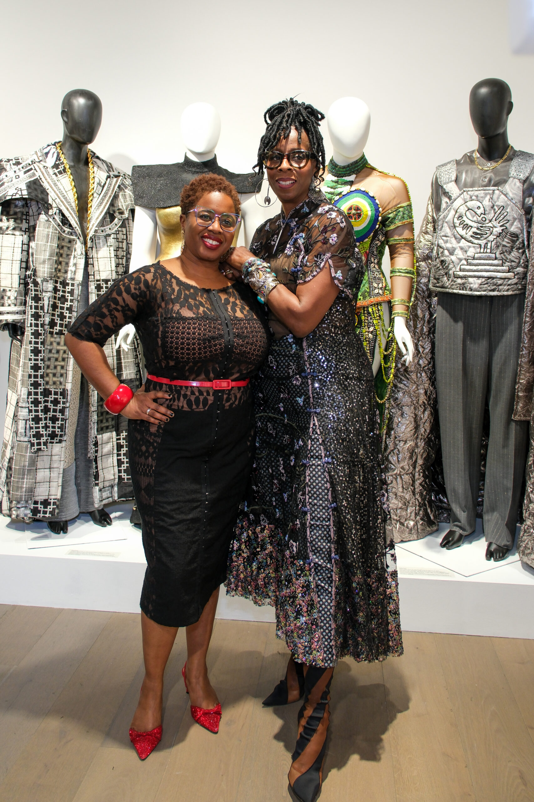Two stylish women pose together at a fashion exhibition, standing in front of mannequins adorned with intricate, avant-garde designs. The woman on the left wears a black lace dress with a red belt and matching red accessories, including a bracelet and pointed heels. The woman on the right wears a shimmering, multicolored sequined dress with sheer details and dramatic lace-up boots. They smile warmly, embracing in a celebratory moment.