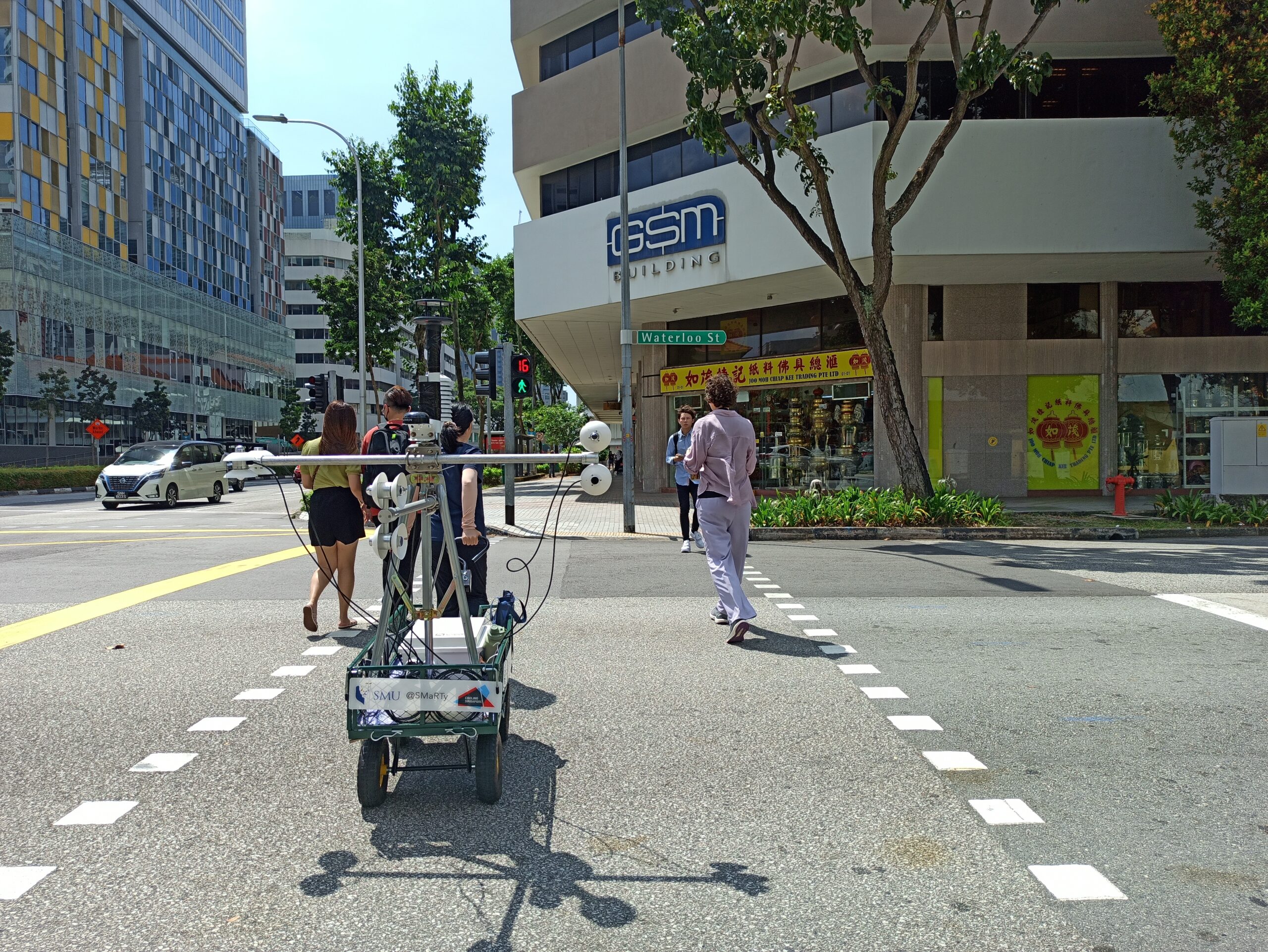 Pedestrians cross the street on a bright, sunny day as a heat measuring device on wheels with long horizontal arms trails behind them