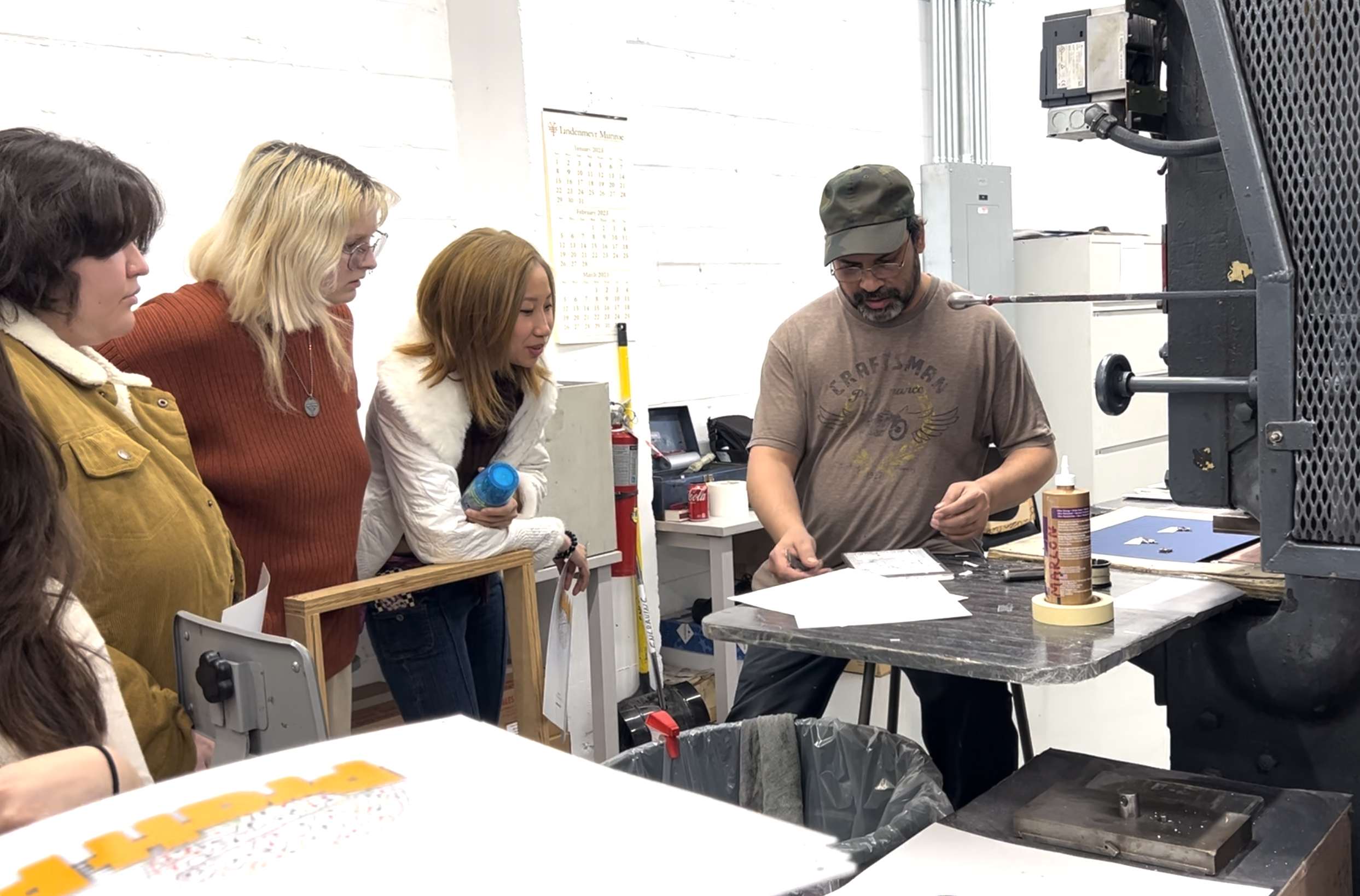 A close-up of the technician in the printing workshop, engaging with a few students as he demonstrates the printing process. One student with blonde hair and glasses, another with a white jacket, and a third in a mustard-colored coat are closely observing and interacting with the printing materials.