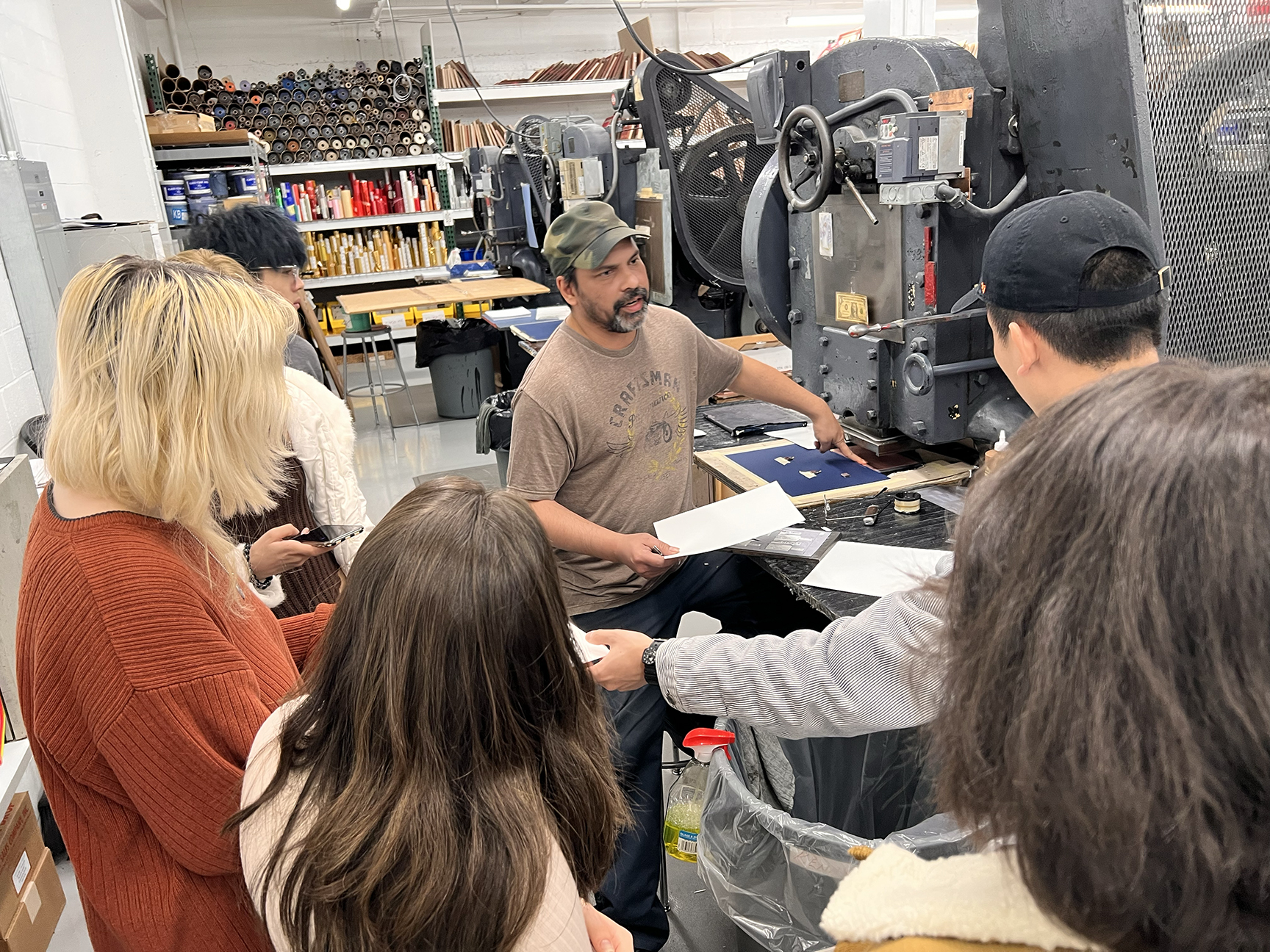 A group of students in a printing workshop attentively observing a technician demonstrating the use of a large industrial printing press. The technician, wearing a brown t-shirt and a green cap, is holding a sheet of paper while explaining the printing process. The background features shelves filled with various printing materials and tools.