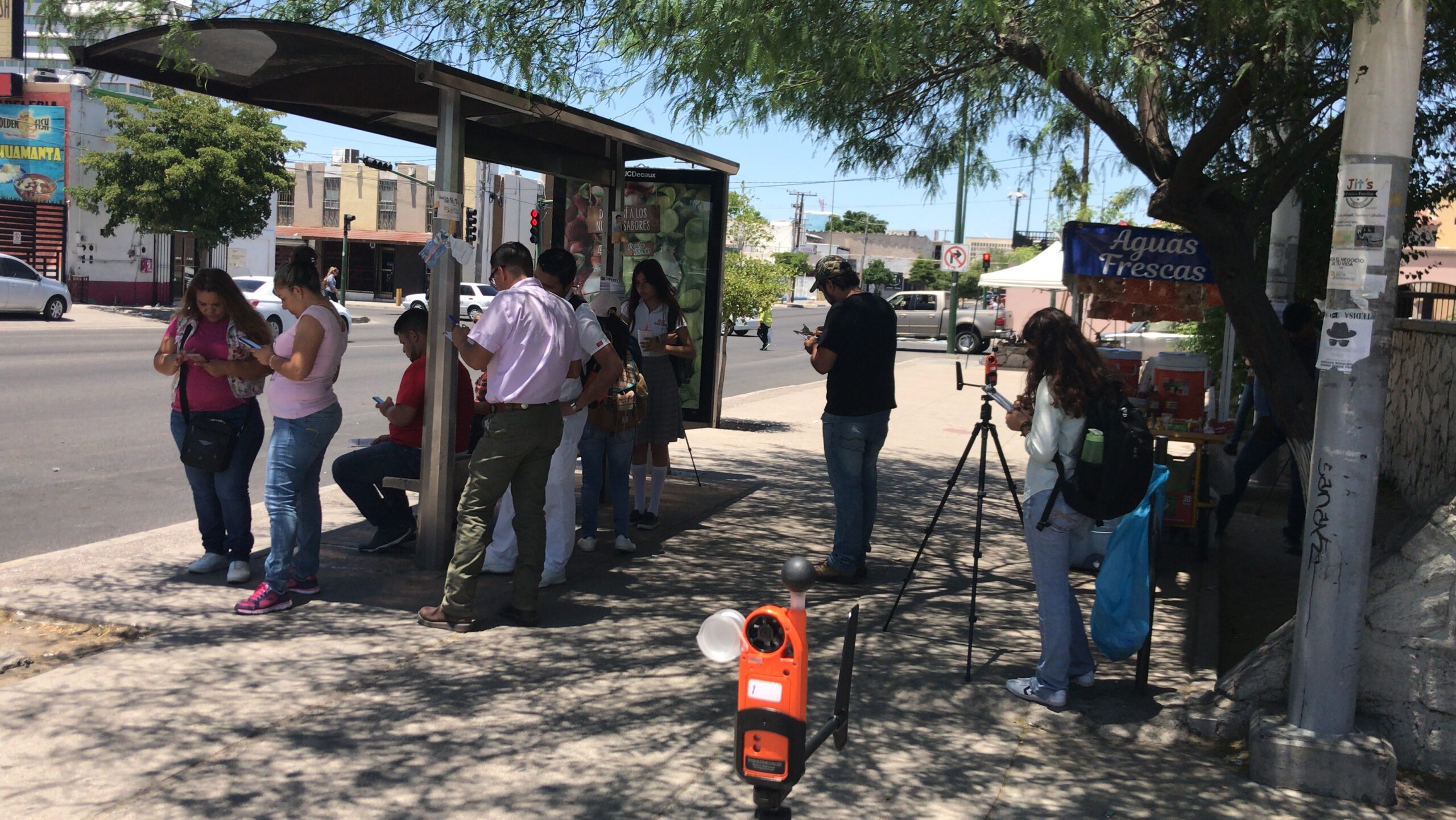A handful of commuters wait in the shade for a bus as someone behind them uses a heat measuring device attached to a tripod