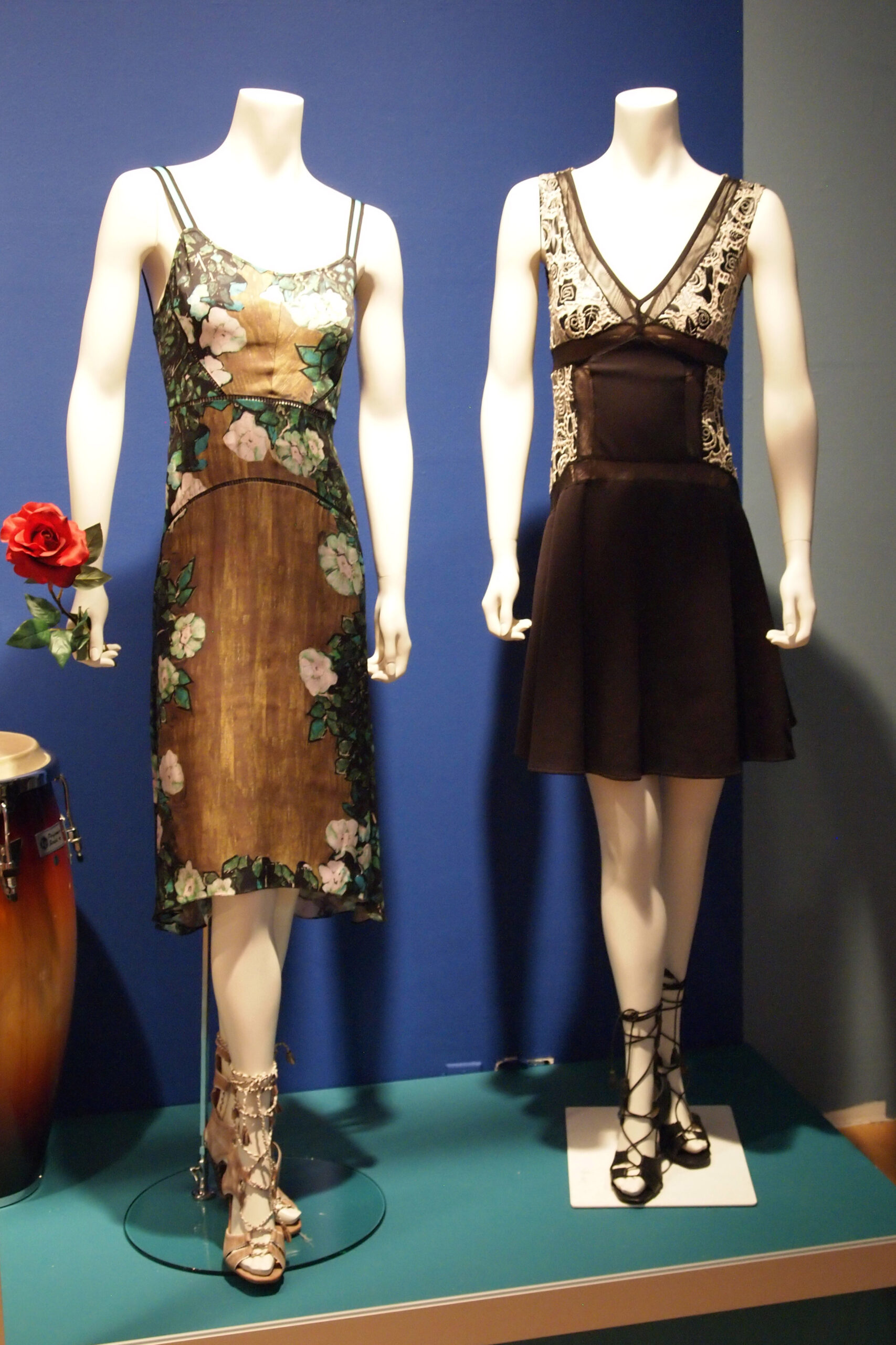 A gallery display featuring two mannequins dressed in elegant Tracy Reese dresses against a blue background. The mannequin on the left wears a midi-length slip dress with a floral print, sheer overlay, thin straps, and intricate detailing, accessorized with a red rose in its hand and strappy heeled sandals. The mannequin on the right is dressed in a black sleeveless dress with a fitted bodice, lace details on the upper portion, a plunging neckline, and a flared skirt, paired with lace-up black heels.