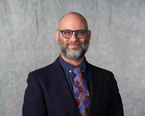 A professional headshot of a man with a bald head, beard, and glasses, wearing a dark suit jacket, navy shirt, and a colorful patterned tie. He is smiling slightly and posed in front of a neutral gray backdrop.