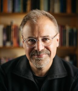 A professional portrait of a man with glasses, a beard, and mustache, smiling softly. He is wearing a dark collared sweater, posed in front of bookshelves filled with books, giving the impression of an academic or writer.