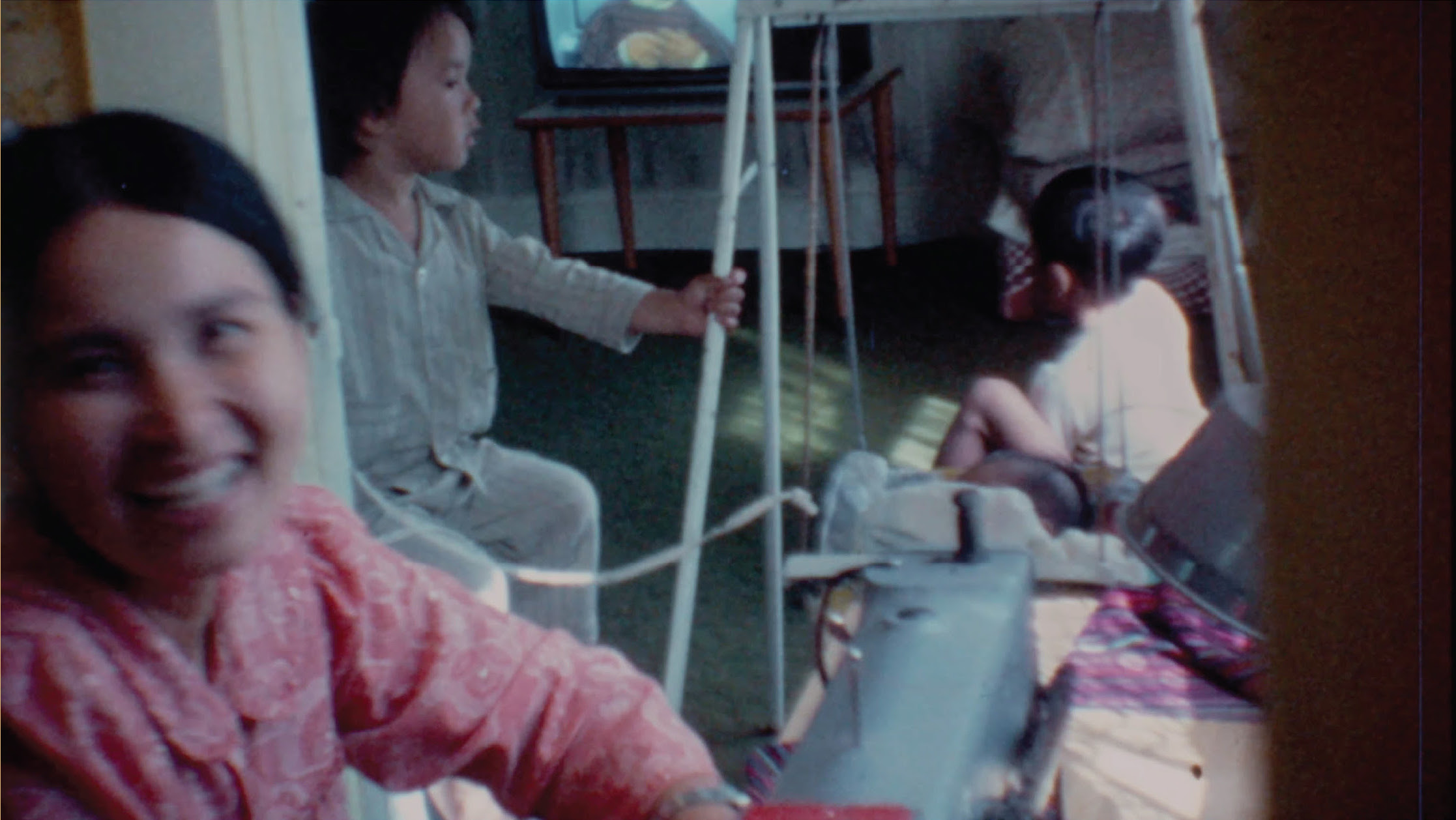 A vintage-style film still depicting a warmly lit domestic scene. In the foreground, a woman in a patterned pink shirt is smiling towards the camera, slightly out of focus. Behind her, a young child wearing pajamas is sitting on the floor, holding onto a swing frame, while another child is seated on the swing, facing away. A television screen in the background displays an animated character, and the room is dimly lit with natural light casting soft shadows.